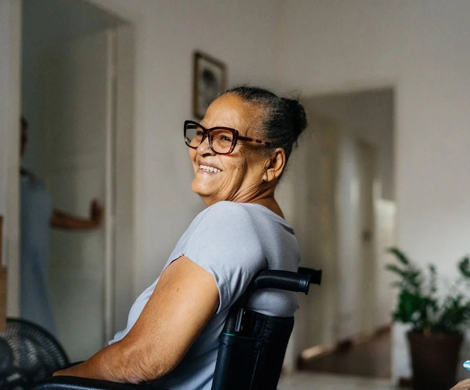 A smiling elderly woman with glasses, sitting in a wheelchair in a cozy, well-lit living room with a plant and artwork in the background.