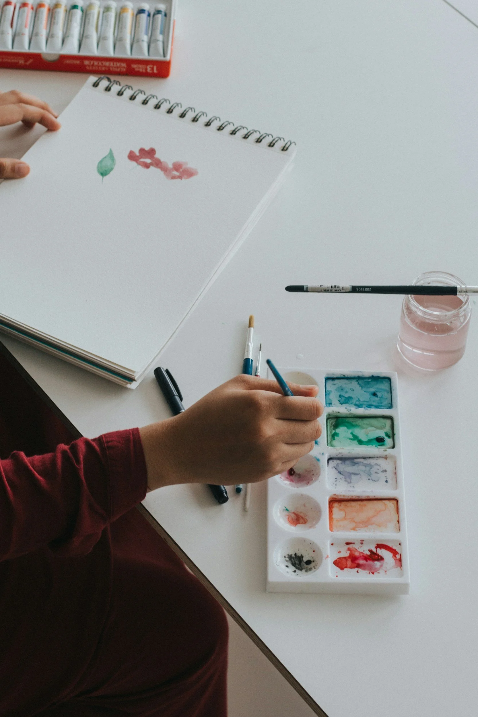 Person painting with watercolor paints on paper, with additional brushes and a palette of colors on a white table.