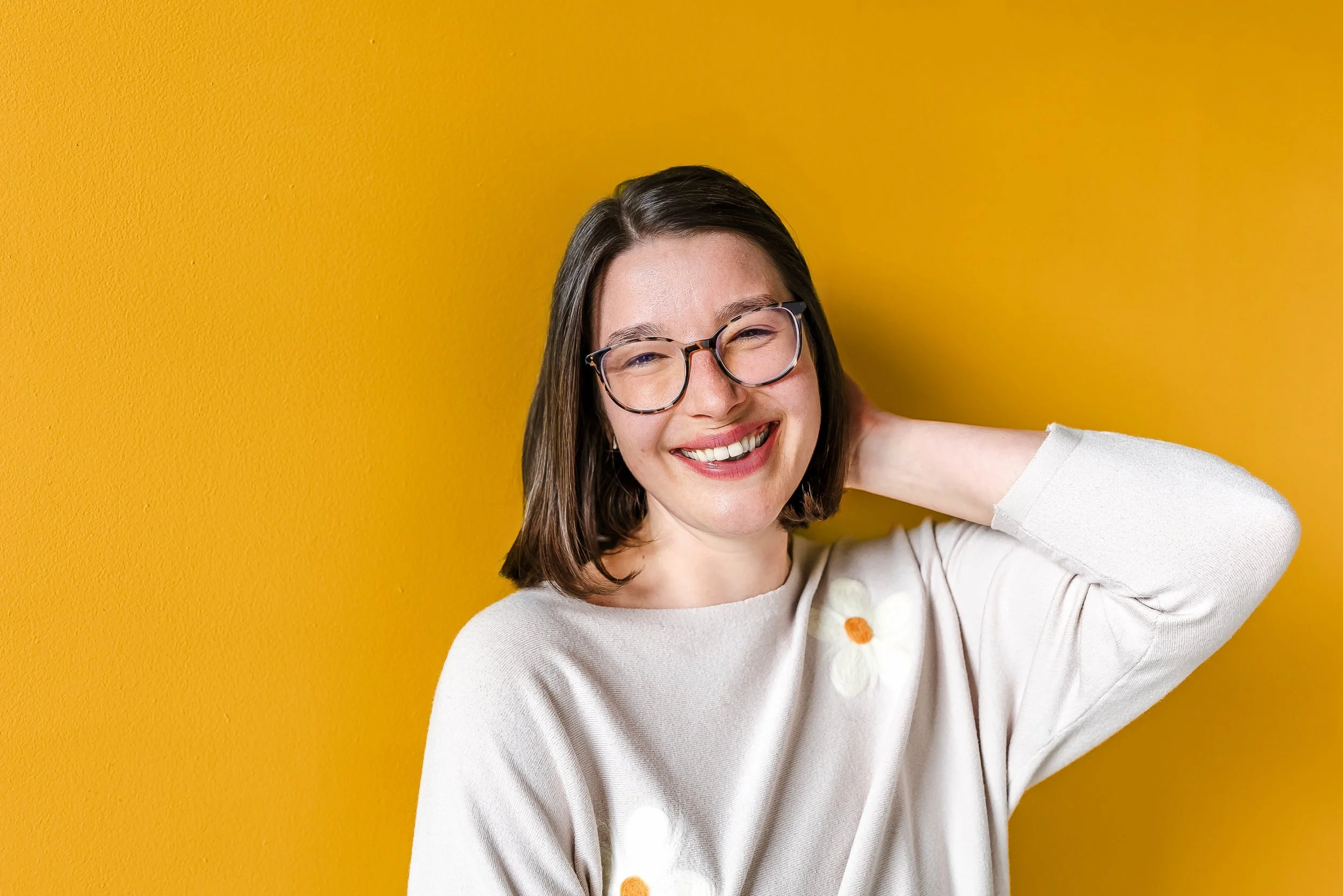 Sarah, an executive function coach, is standing in front of a yellow background. She is wearing a tan sweater with white flower and is tucking her hair behind her ear while smiling