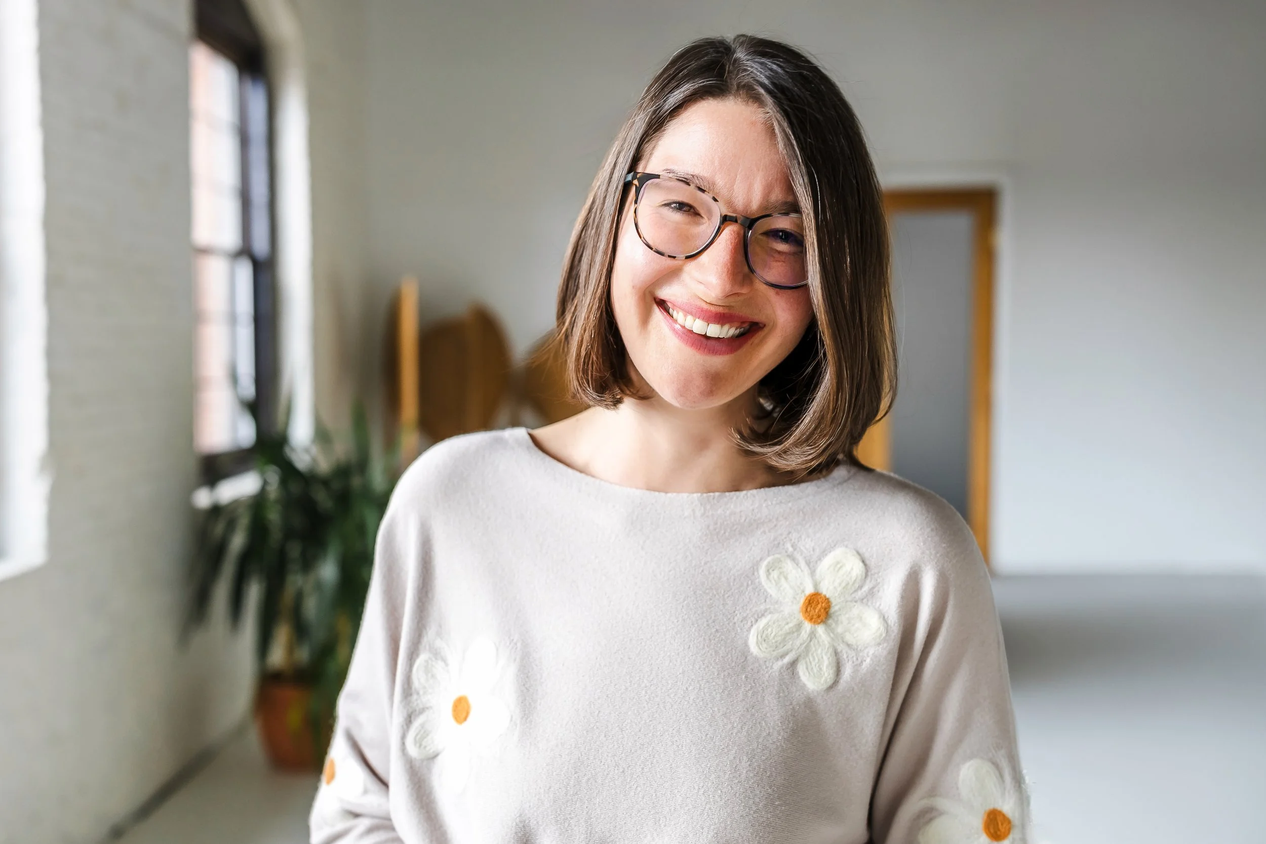 Sarah, an executive function coach, is wearing a tan sweater with white flowers, smiling, wearing glasses