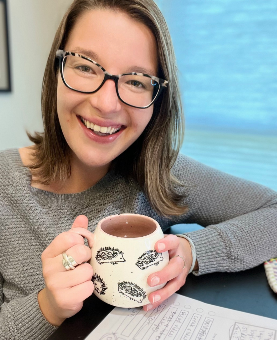 A woman with glasses smiling while holding a ceramic mug with hedgehog illustrations, sitting at a table with papers in front of her.