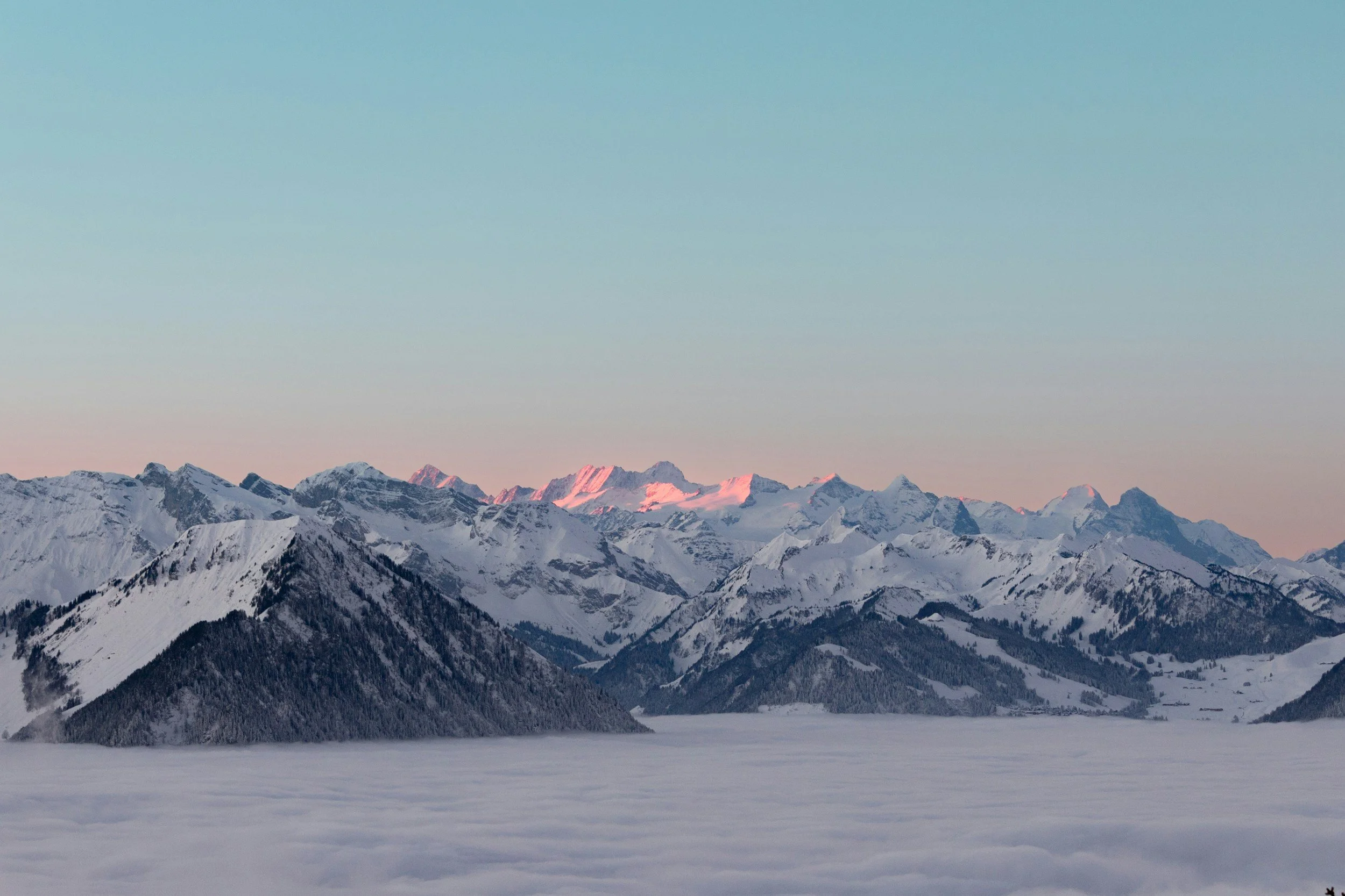 Snow-covered mountain range at sunrise with clouds below and pink hue on mountain peaks.