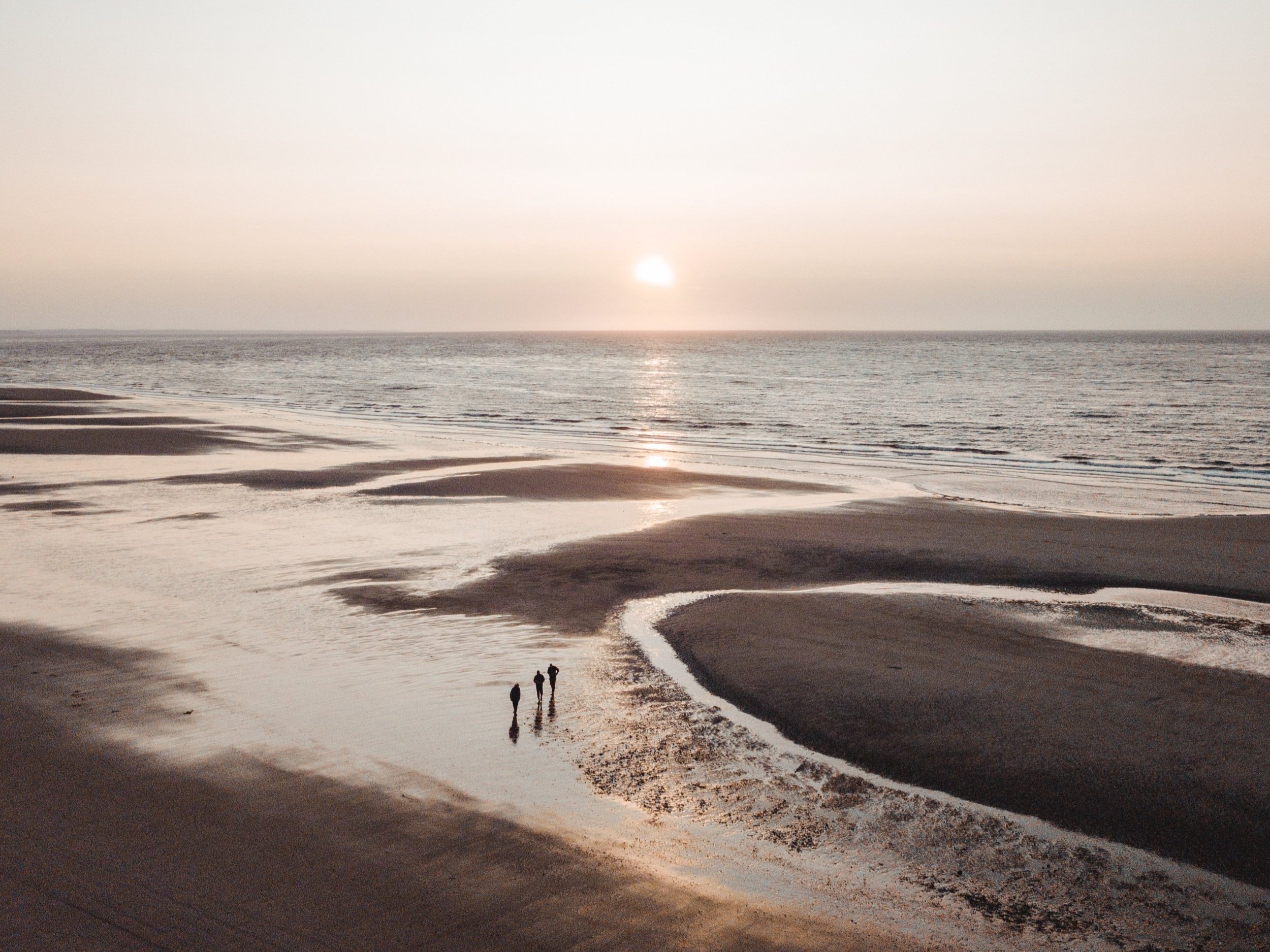 A beach at sunset with three people walking along the wet sand near the water, and water channels creating patterns across the sand.