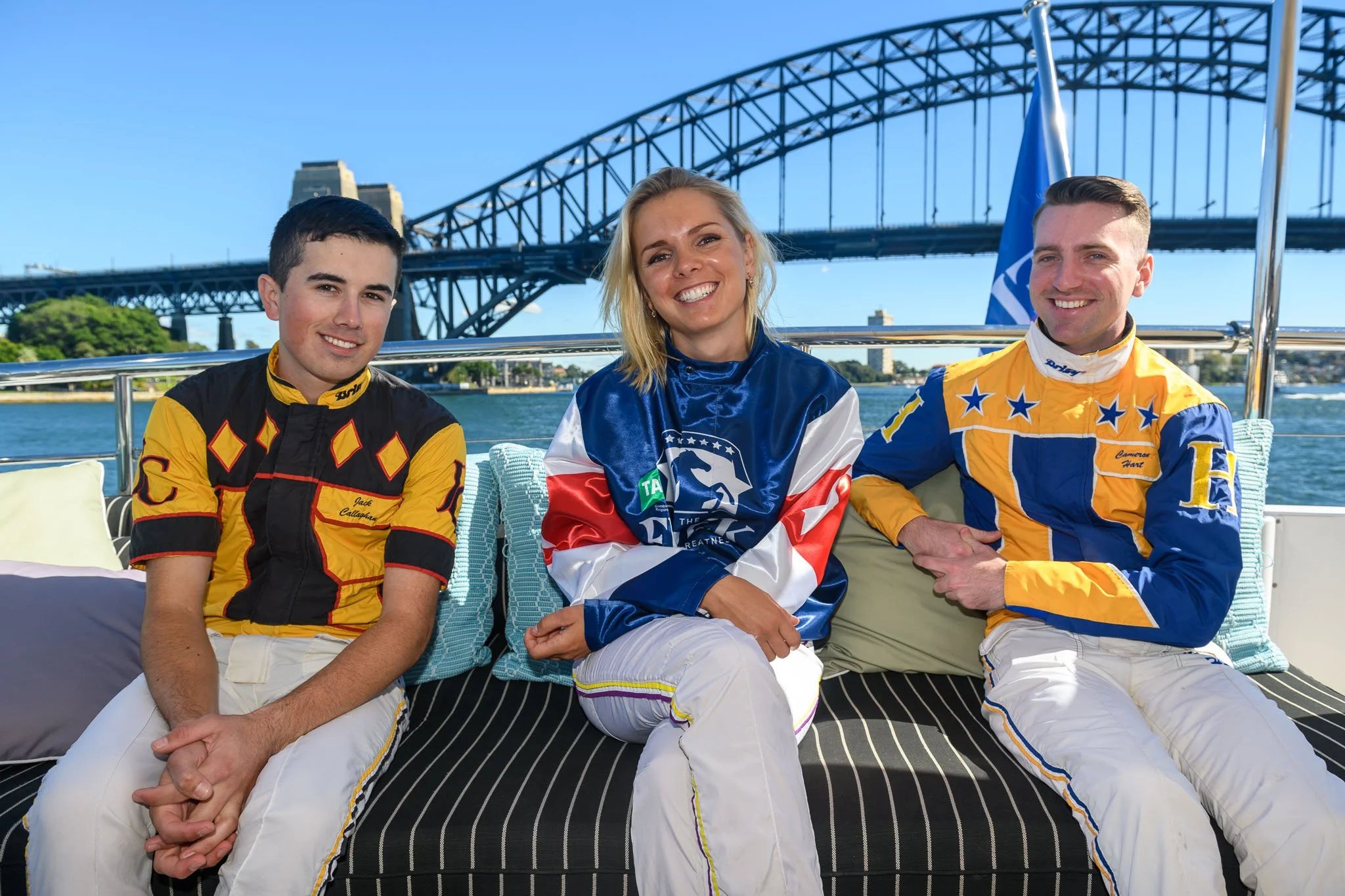 Three race car drivers sitting on a boat with Sydney Harbour Bridge in the background, wearing racing suits and smiling.
