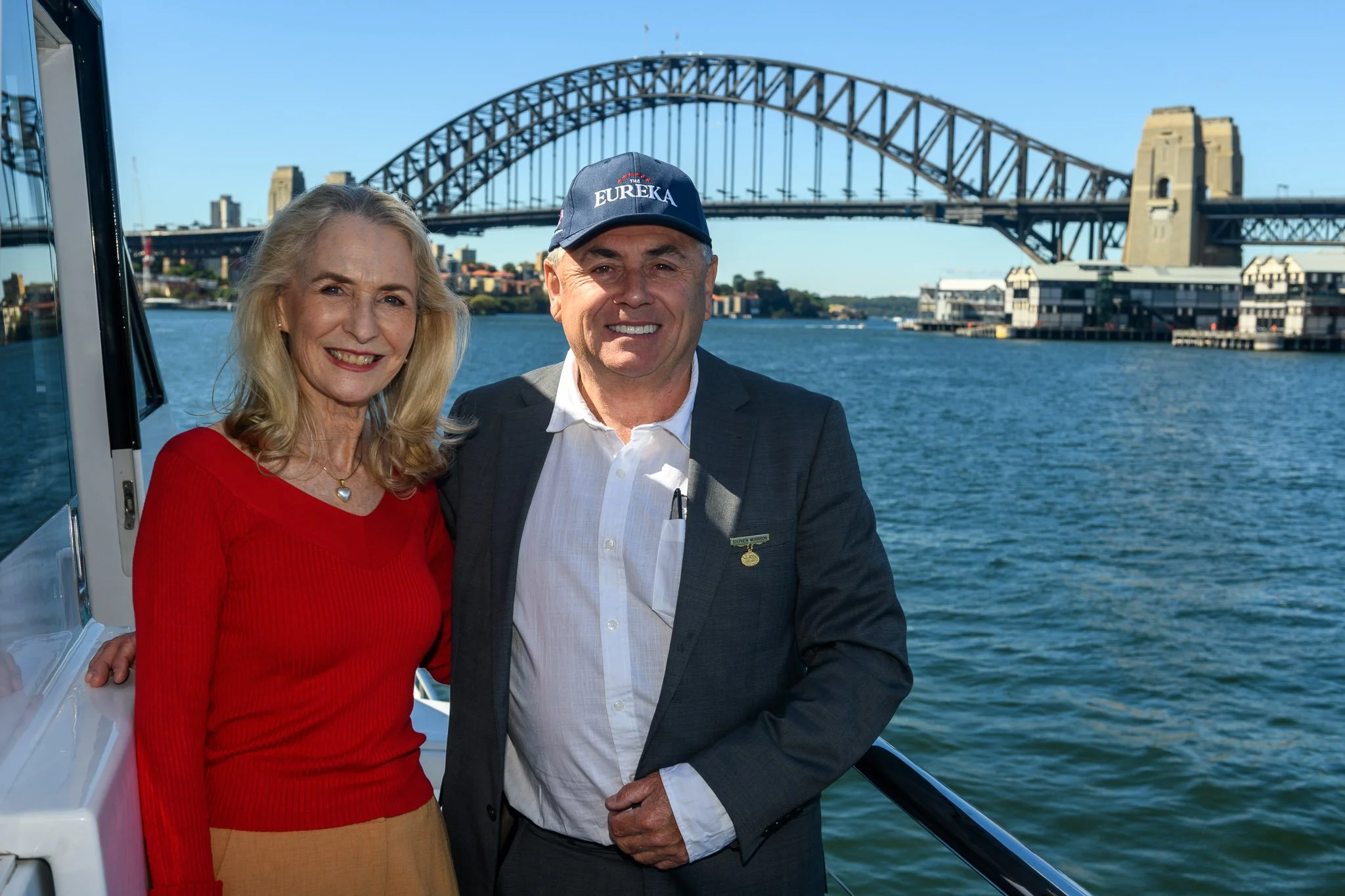A smiling woman in a red sweater and a man in a grey suit with a cap that says 'Eureka' standing on a boat, with the Sydney Harbour Bridge in the background.
