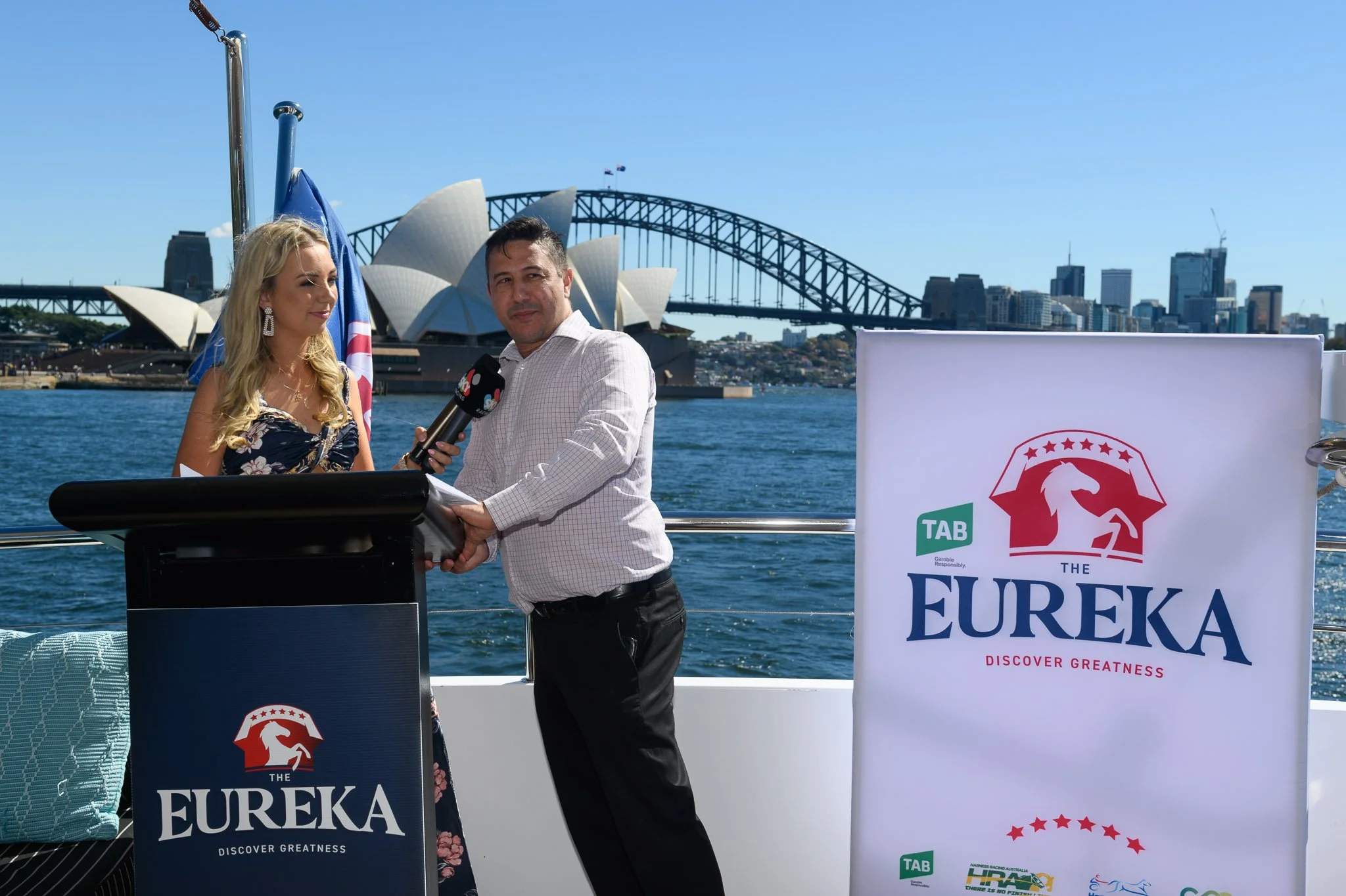 A man and woman standing on a boat during a live interview with Sydney Opera House and Harbour Bridge in the background. The woman holds a microphone, and there is a Eureka-themed banner nearby.