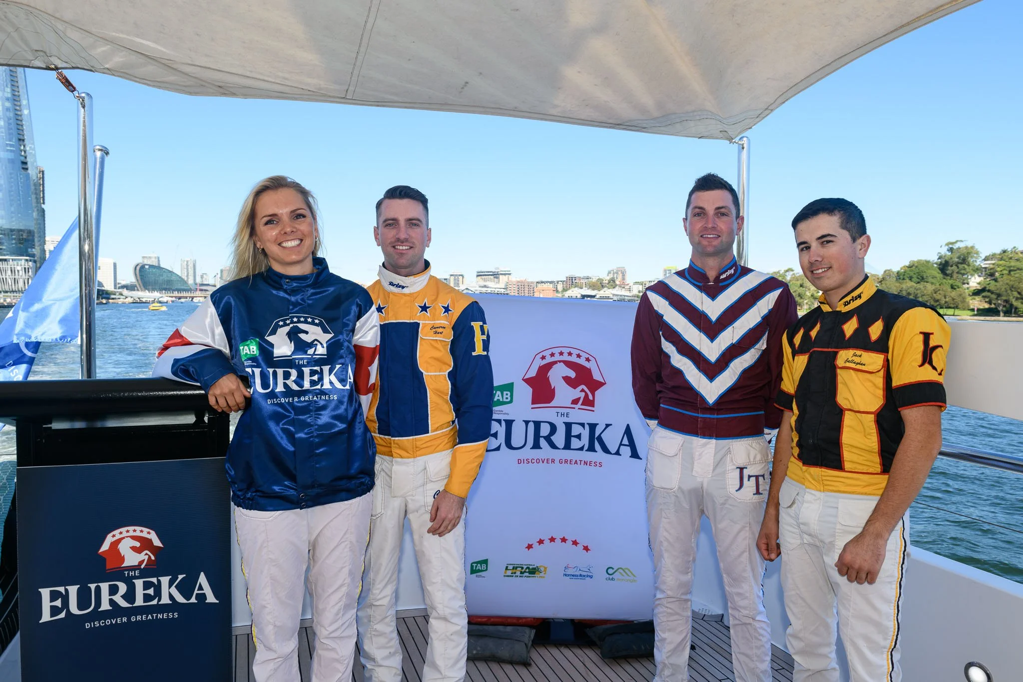 Group of five individuals dressed in racing uniforms standing on a boat with water and cityscape in background, posing in front of a Eureka banner.