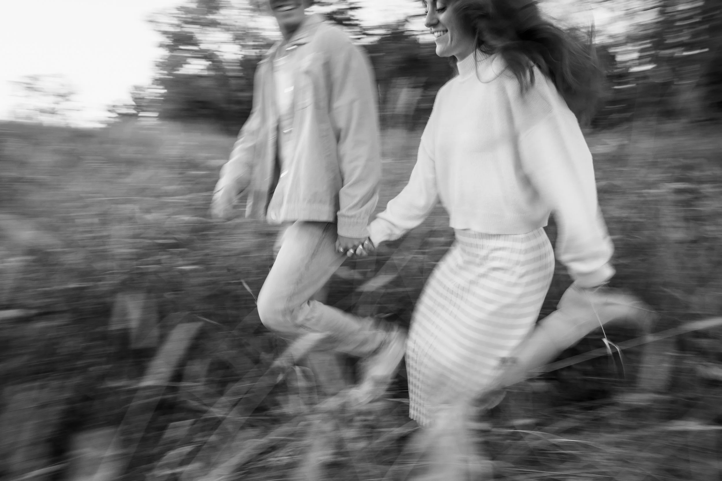 A black-and-white photo of a couple holding hands and running through a field, smiling and enjoying each other's company.
