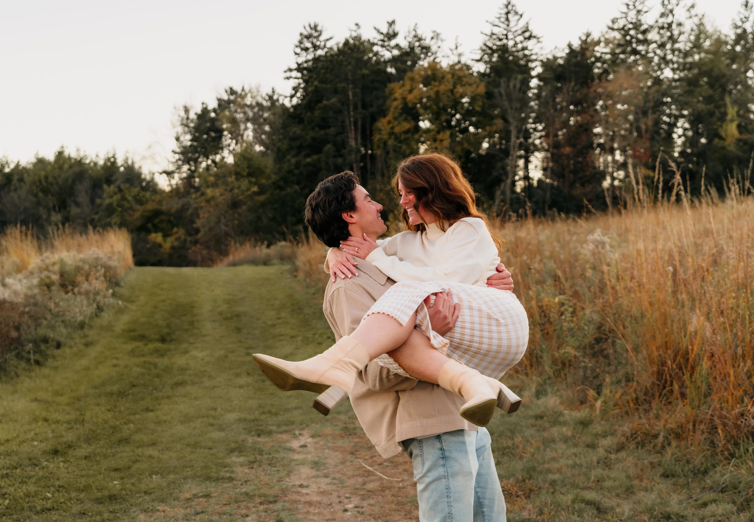 A couple outdoors, man carrying woman in his arms, smiling at each other, in a natural setting with trees and tall grass in the background.