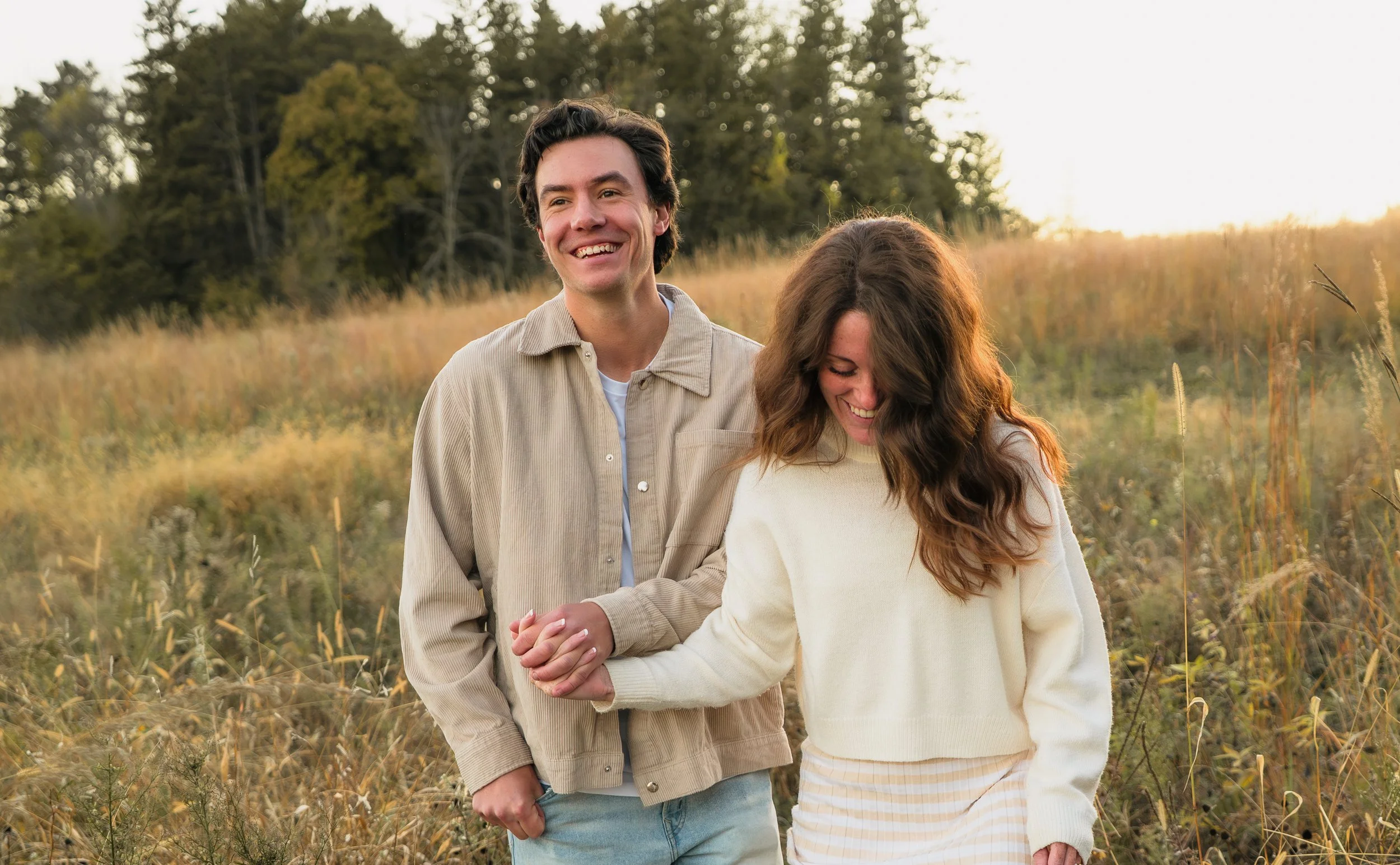 A young couple walking in a field at sunset, holding hands, smiling, with trees in the background.