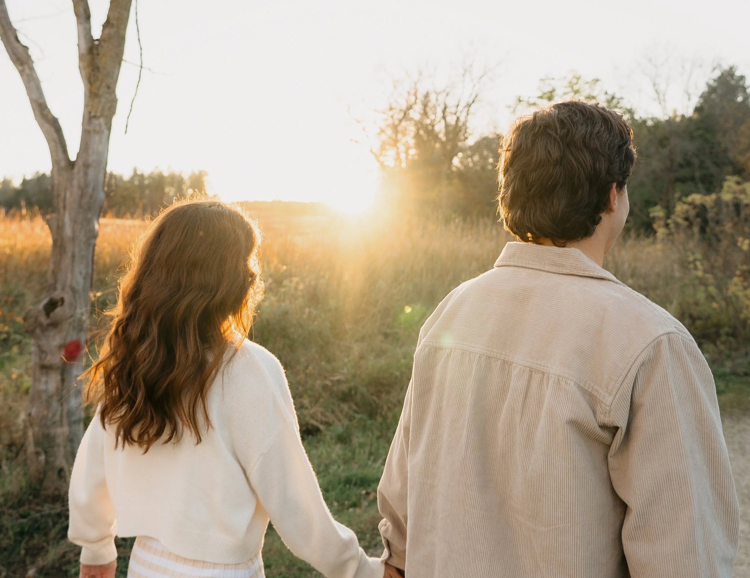 A couple walking outdoors during sunset, holding hands, with trees and tall grass in the background.
