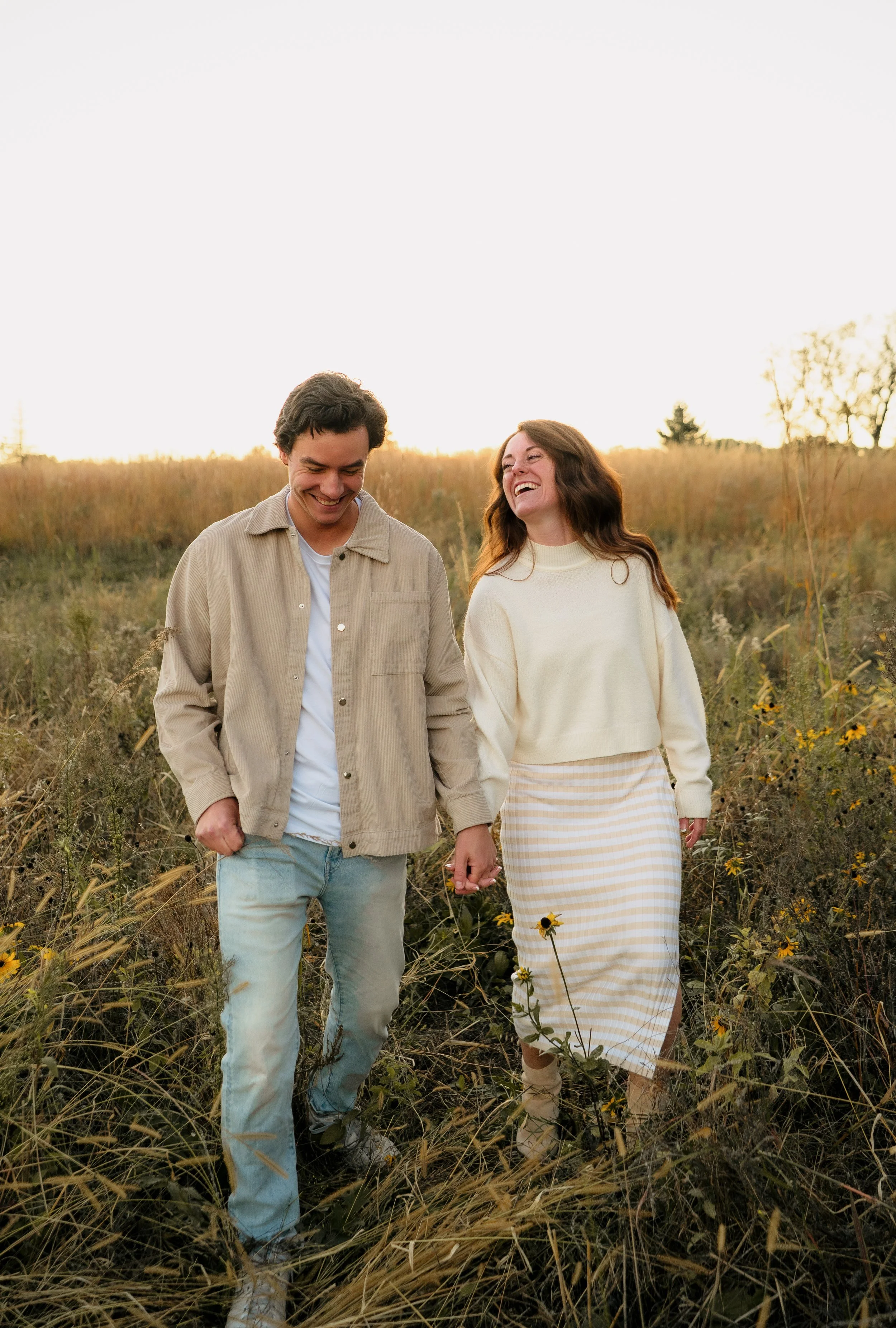 A young couple walking through a grassy field at sunset, holding hands and smiling, dressed in casual clothing.