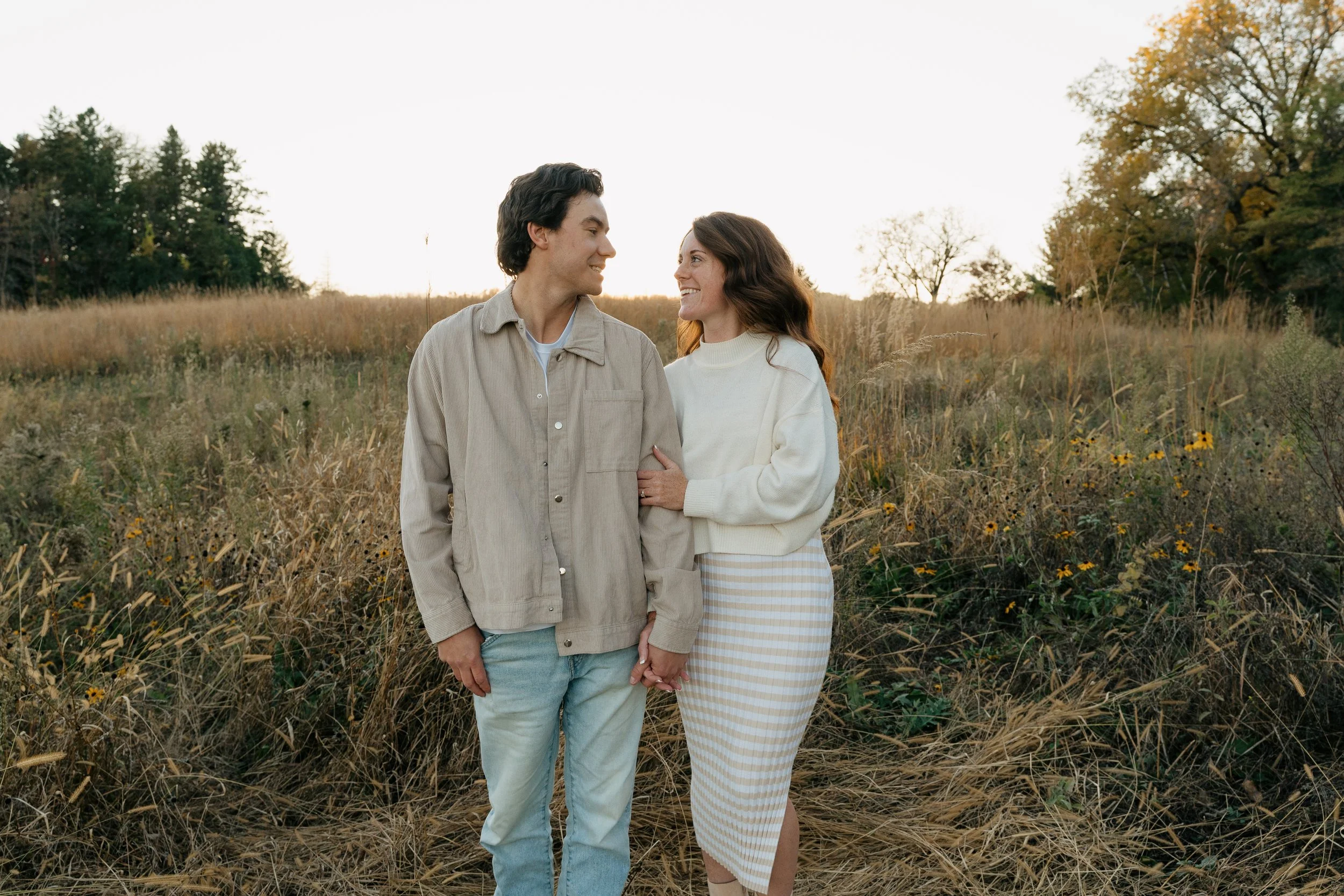A young couple holding hands and smiling at each other in a field during sunset.