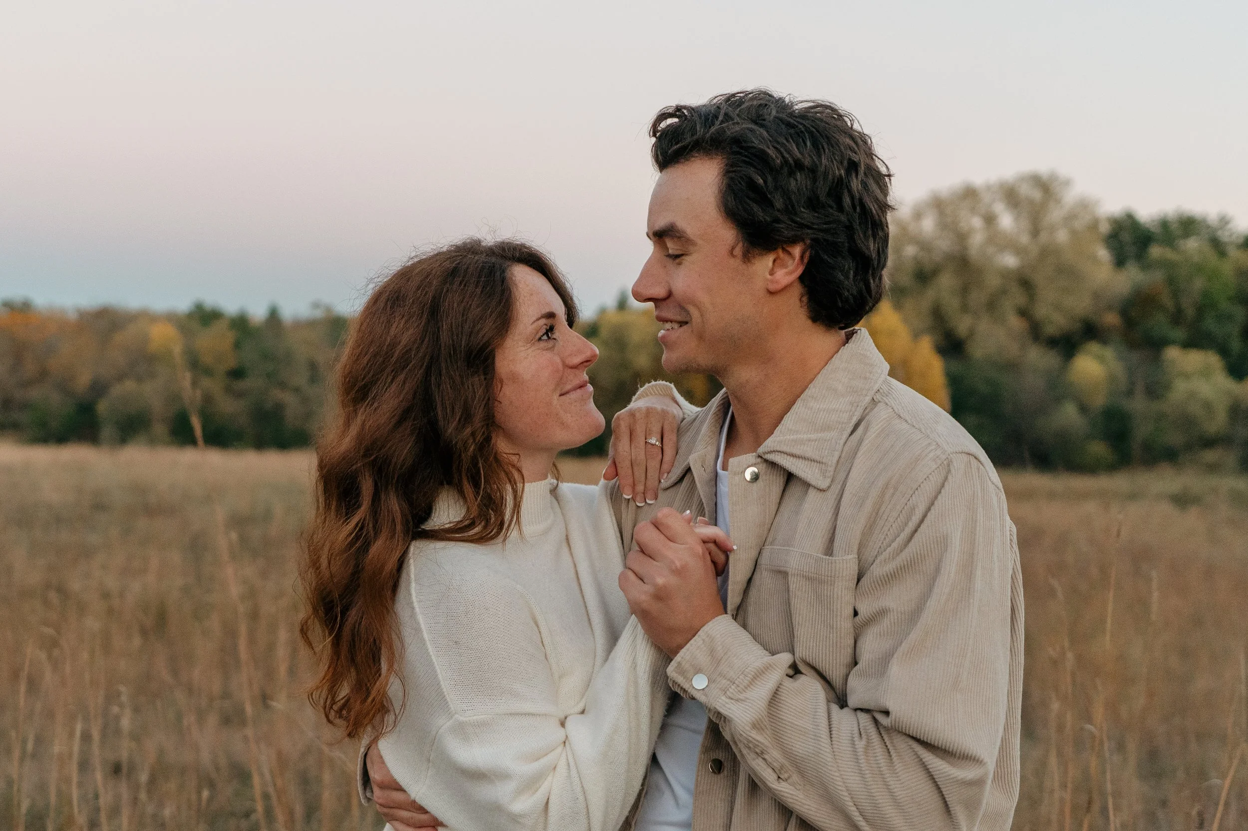 A couple stands in a field during sunset, gazing into each other's eyes, with trees in the background.