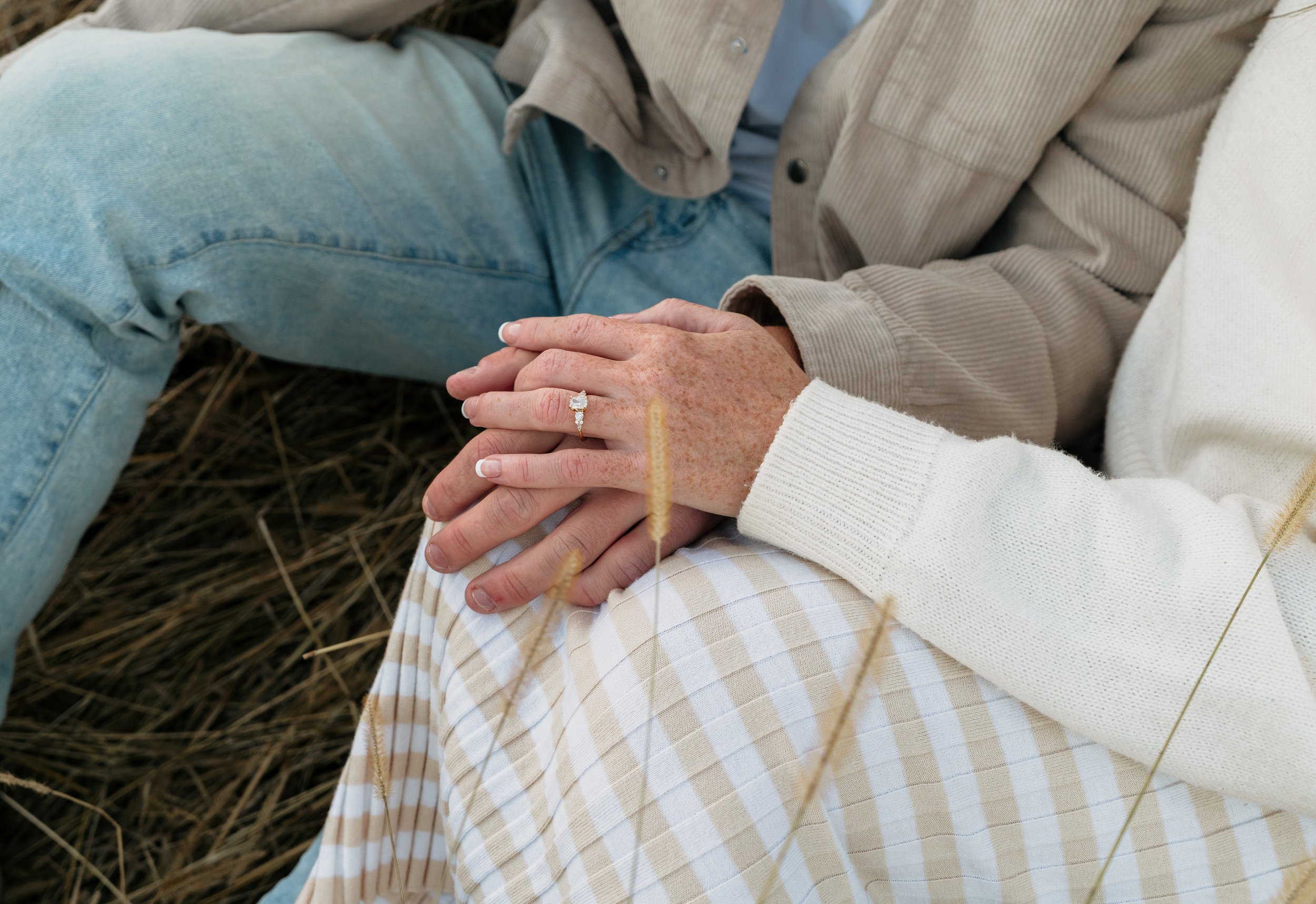 Close-up of two people holding hands, one with a diamond engagement ring, sitting outdoors on grass with dry blades, dressed in light-colored clothing.