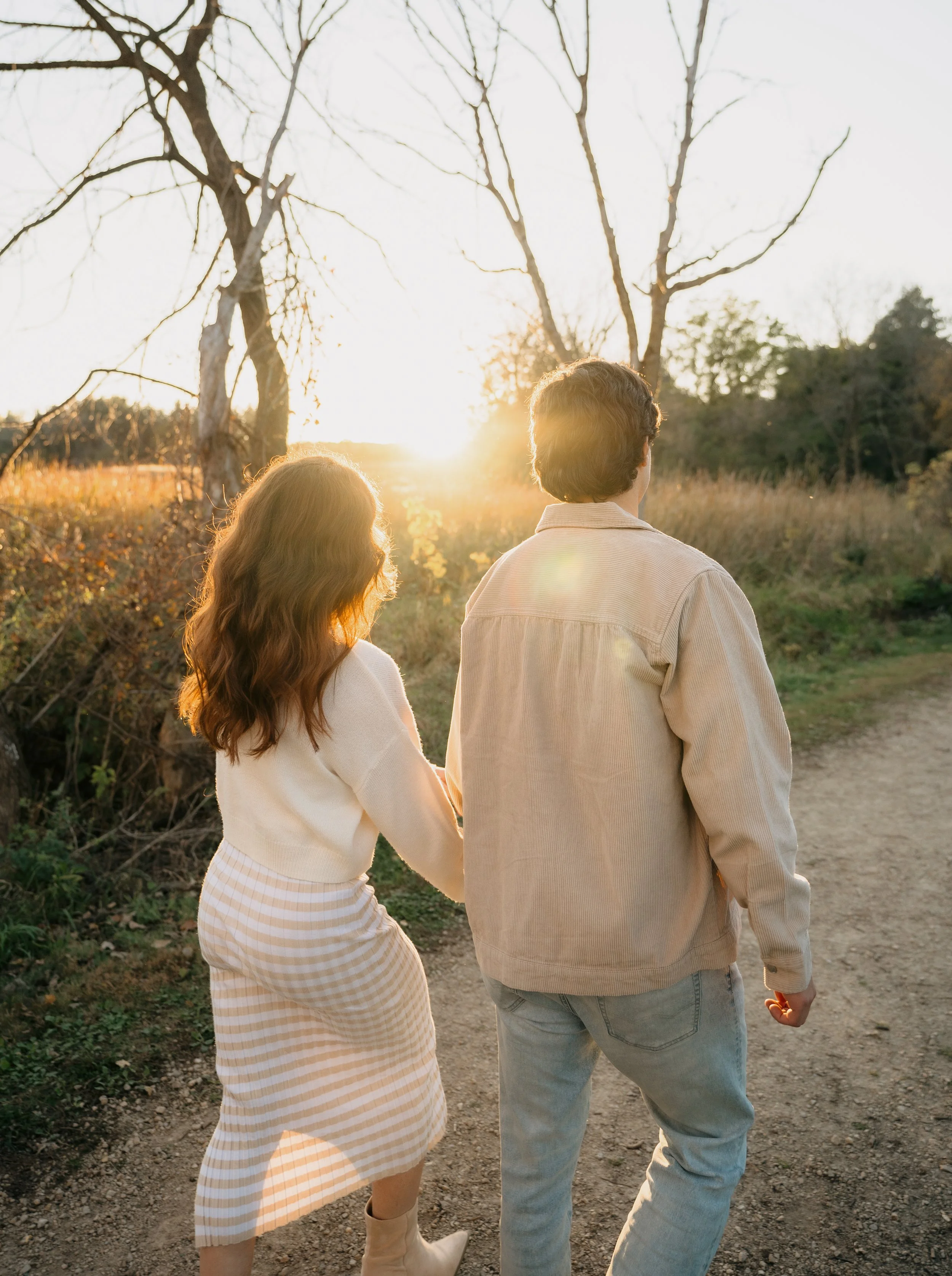 A couple walking hand in hand on a dirt trail during sunset in a rural, wooded area.
