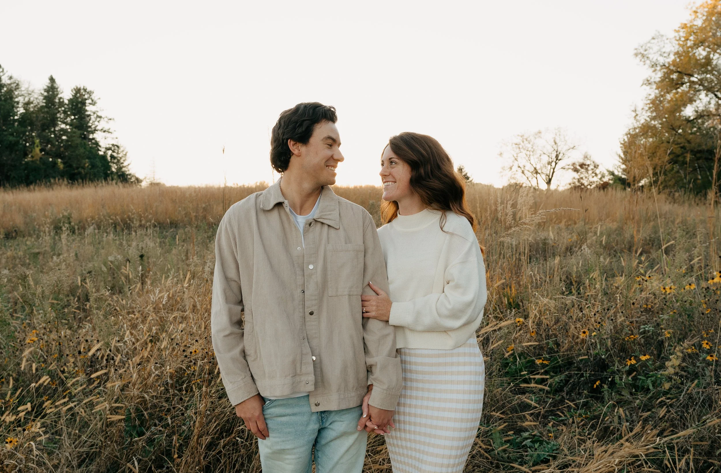A couple holding hands and smiling at each other in a field during autumn.