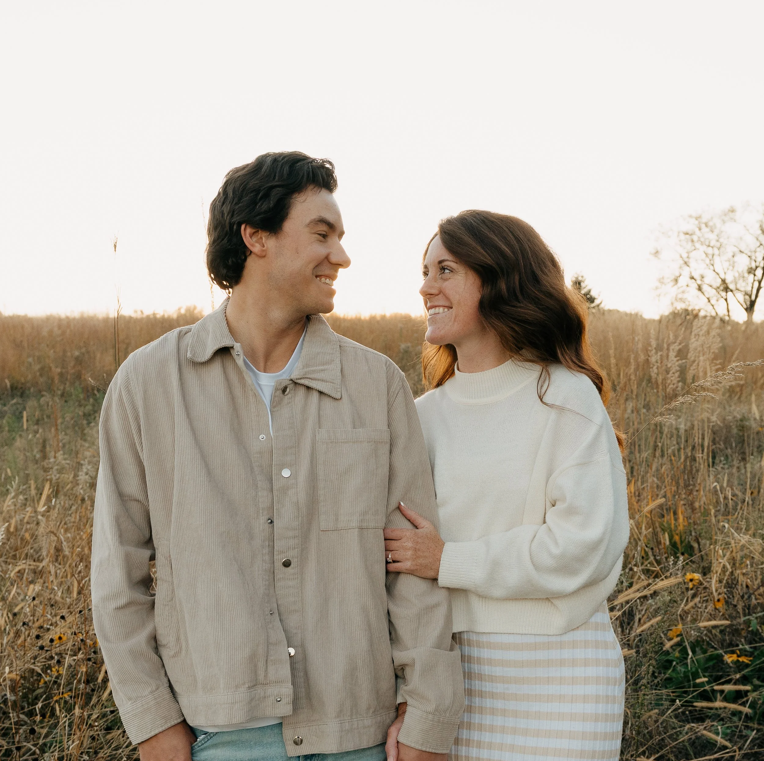 A smiling couple standing in a field of tall grass at sunset, looking at each other.