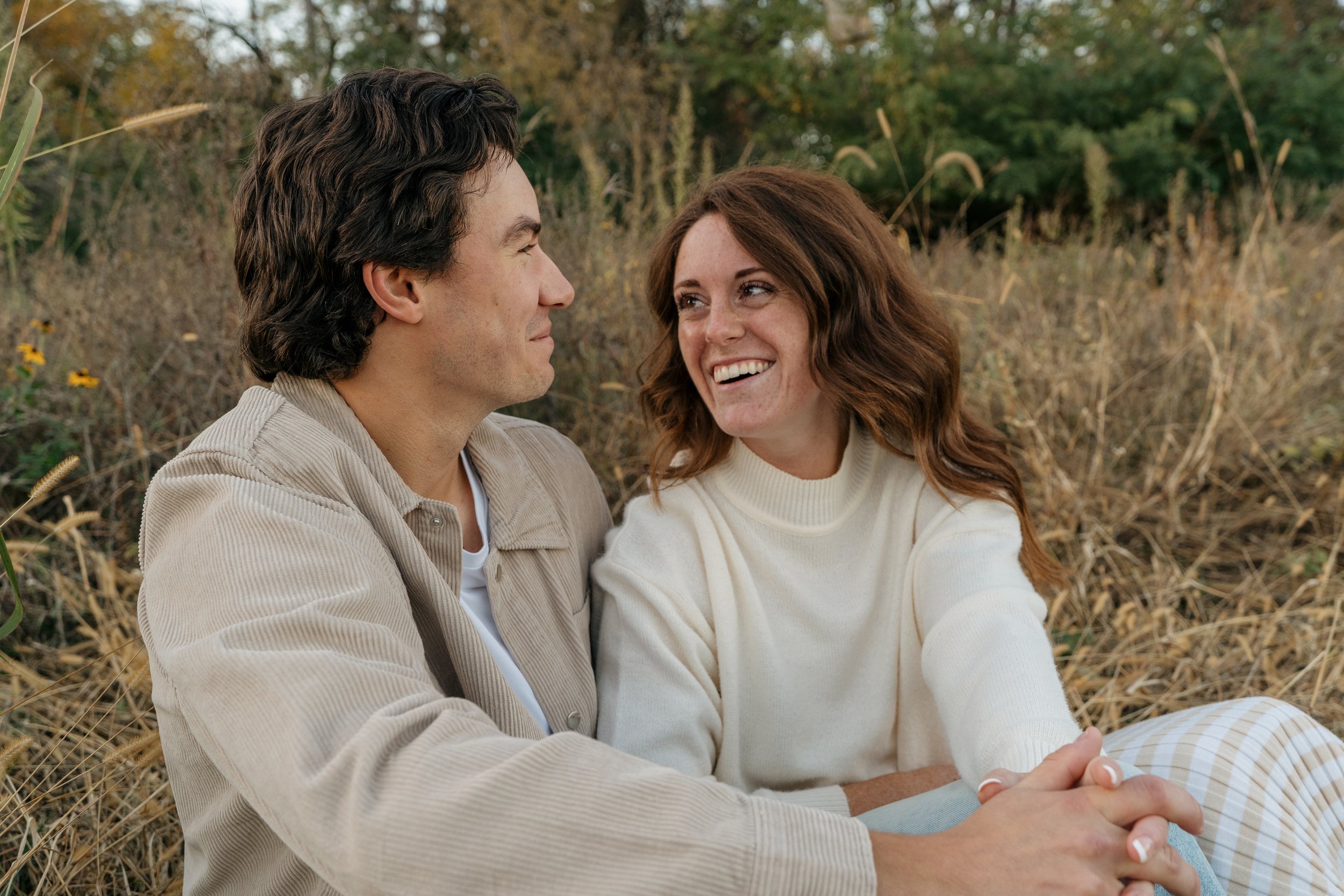 Couple sitting outdoors in a field, smiling and holding hands, with autumn foliage in the background.