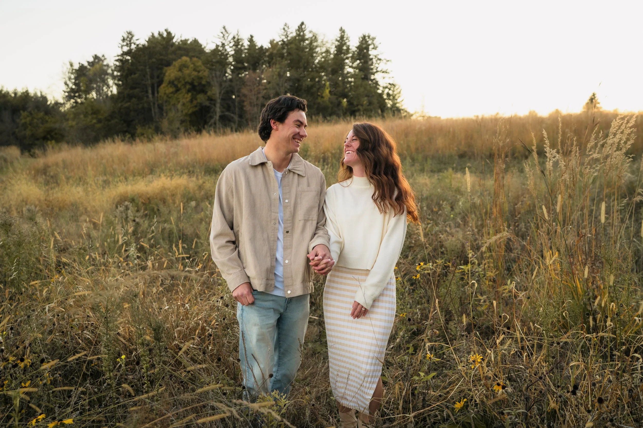 A young couple holding hands and smiling while walking through a grassy field with trees in the background at sunset.