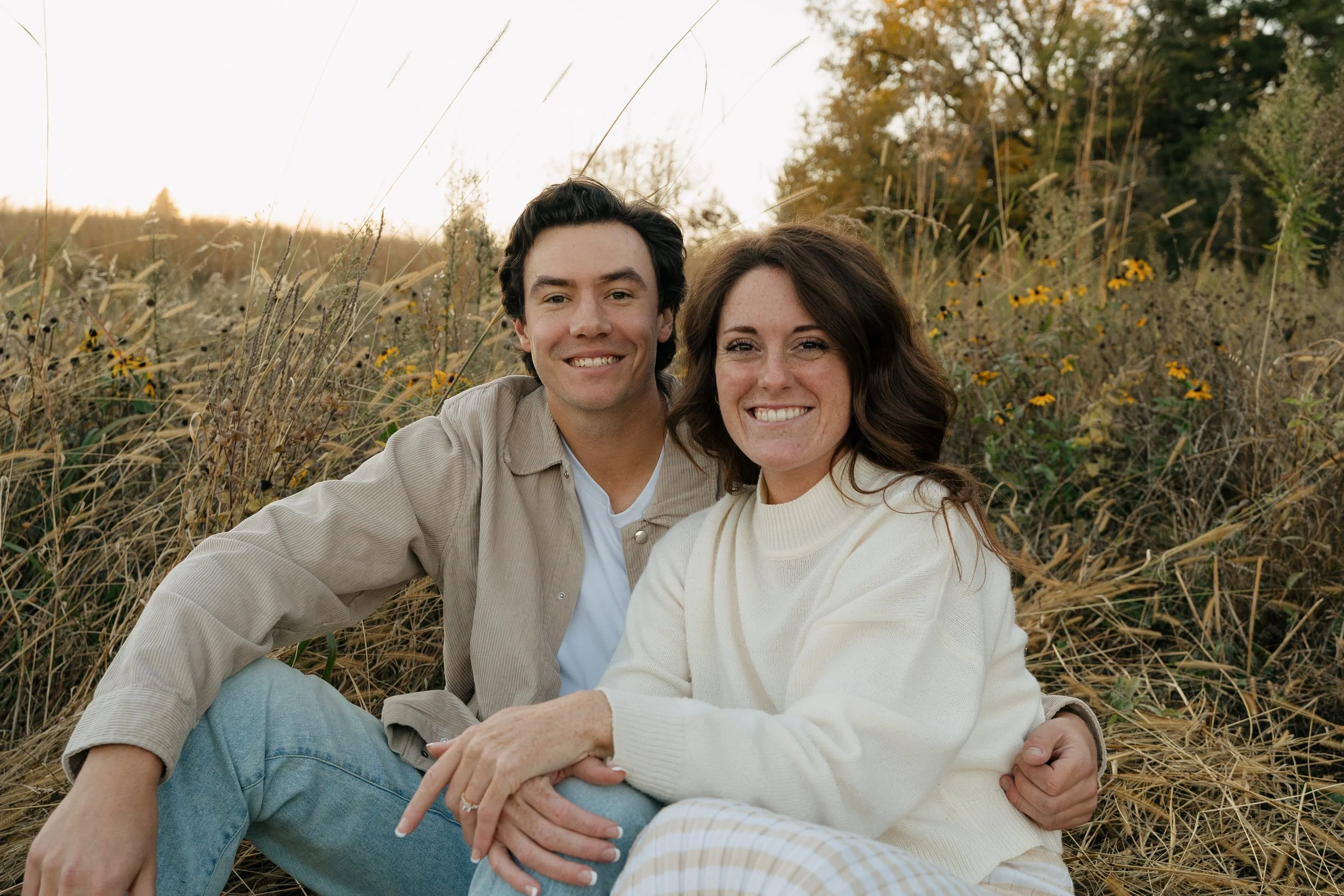 A young man and woman sitting together outdoors in a field of tall grass and yellow flowers, smiling at the camera during sunset.