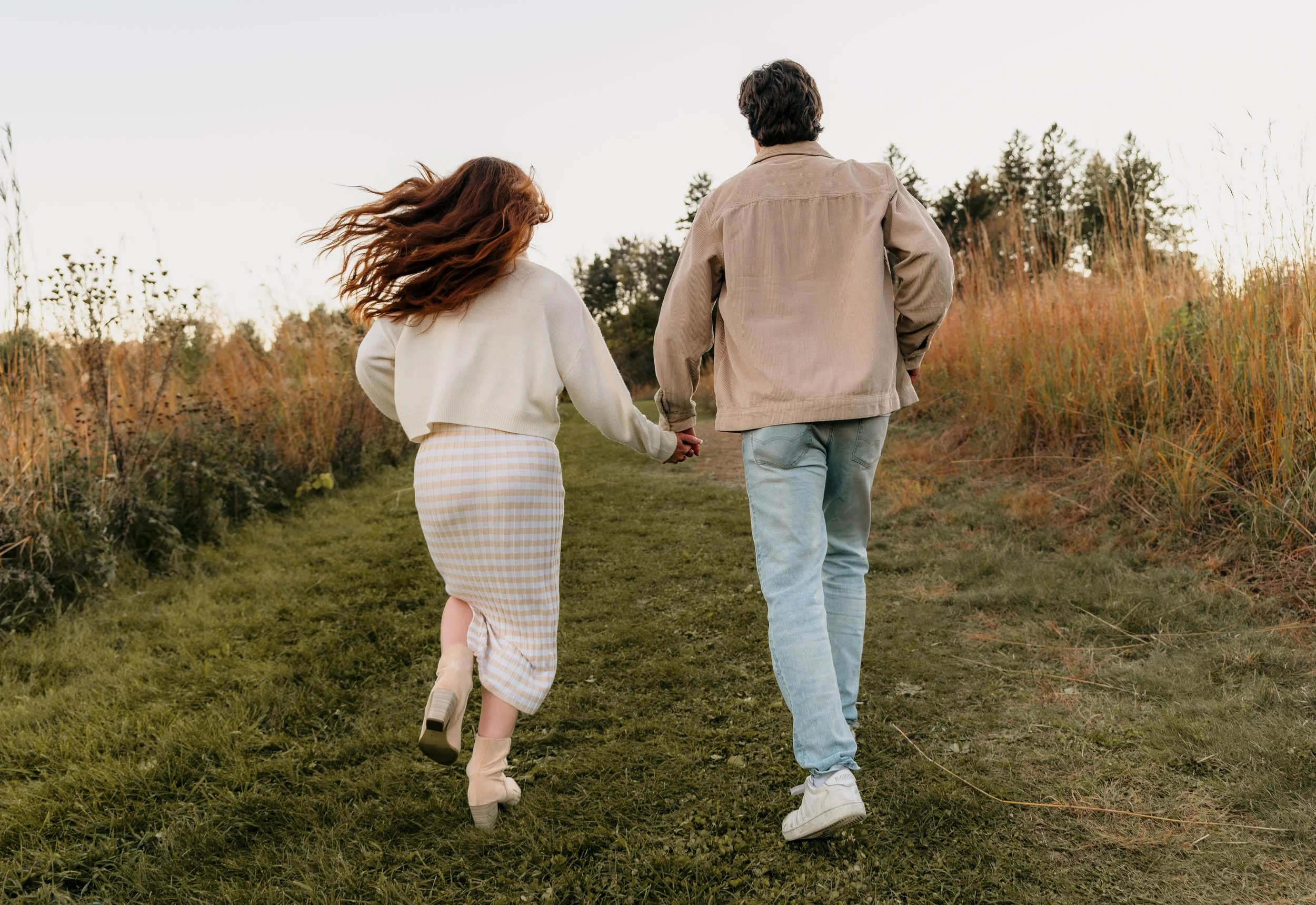 A couple holding hands walking on a grassy trail in a park during fall, with trees in the background.