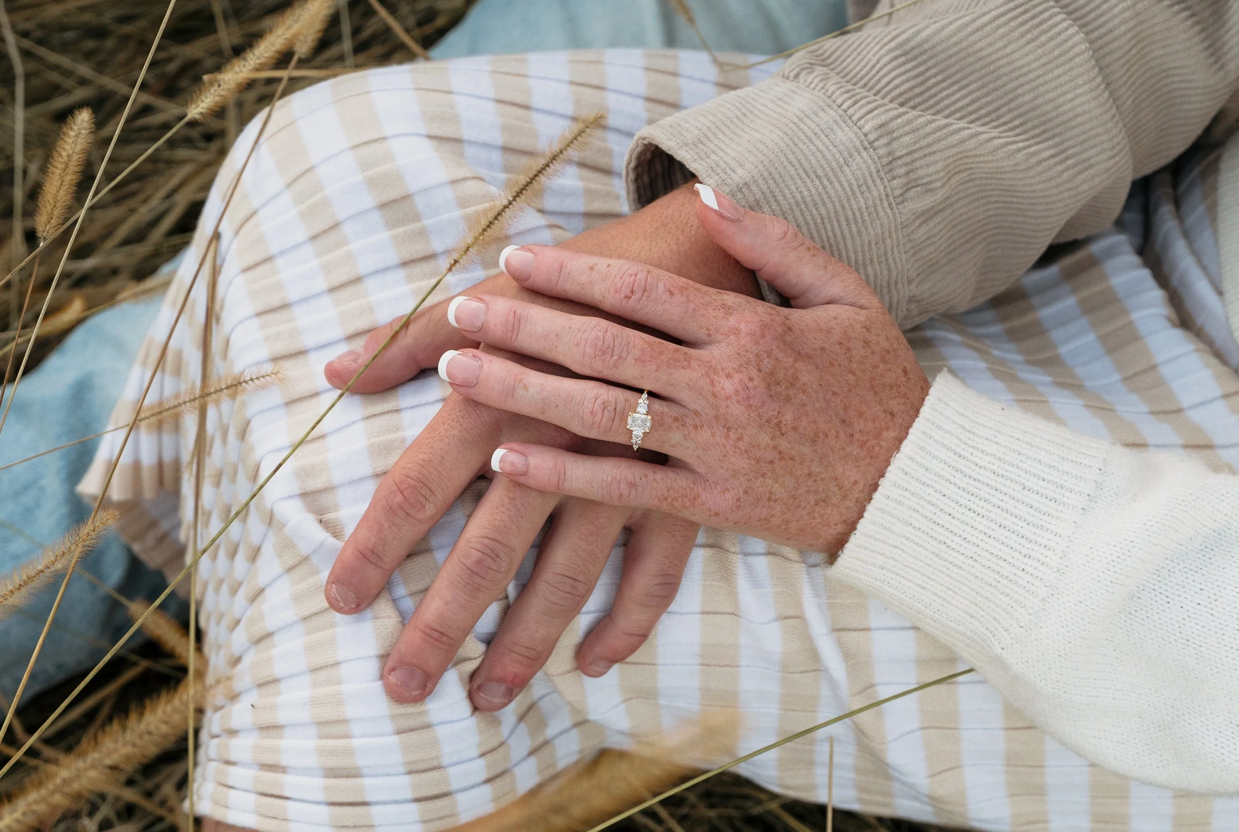 Close-up of two hands gently resting on a lap, one wearing a diamond engagement ring, with the person's arm extended and wearing a cream-colored sweater, in an outdoor setting with dried grass.