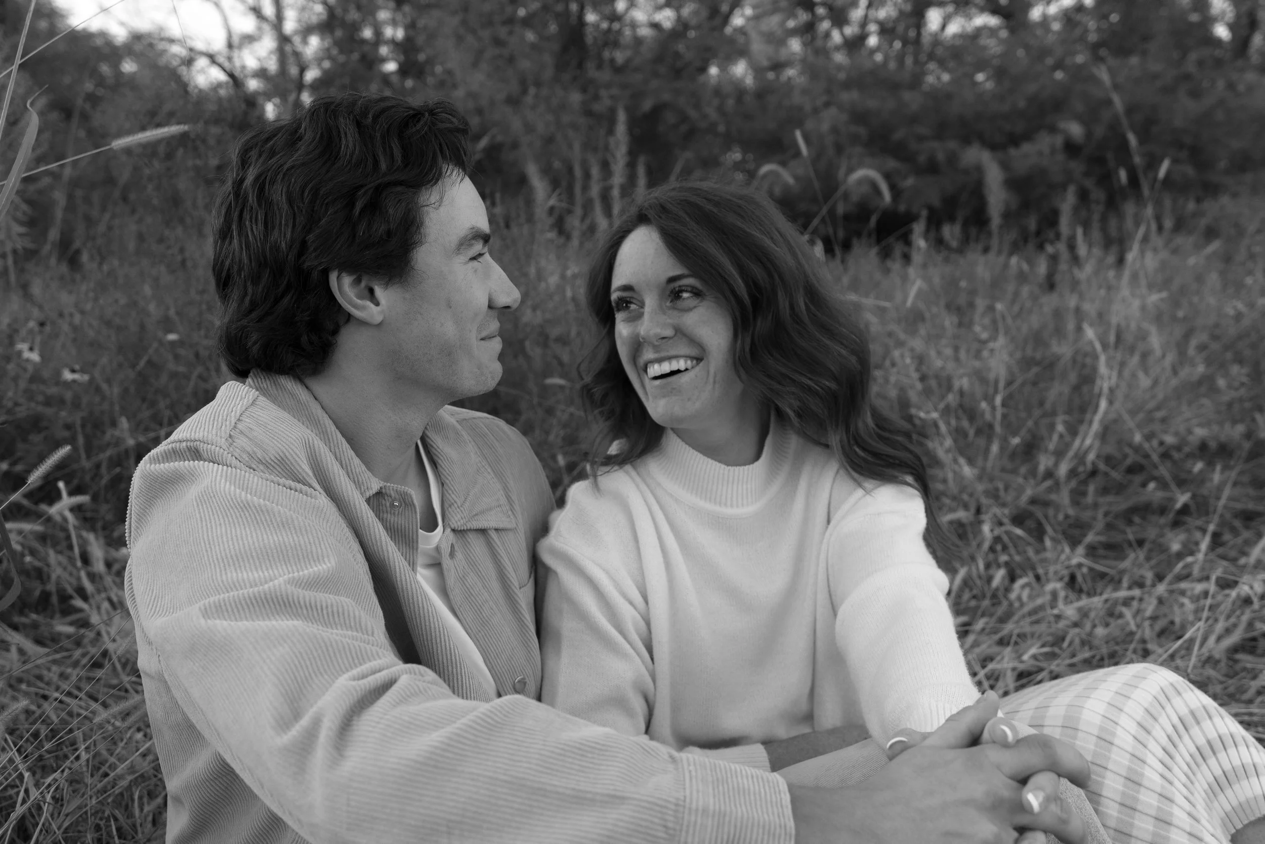 A black and white photo of a smiling woman and a man sitting closely outdoors on grass, looking at each other.