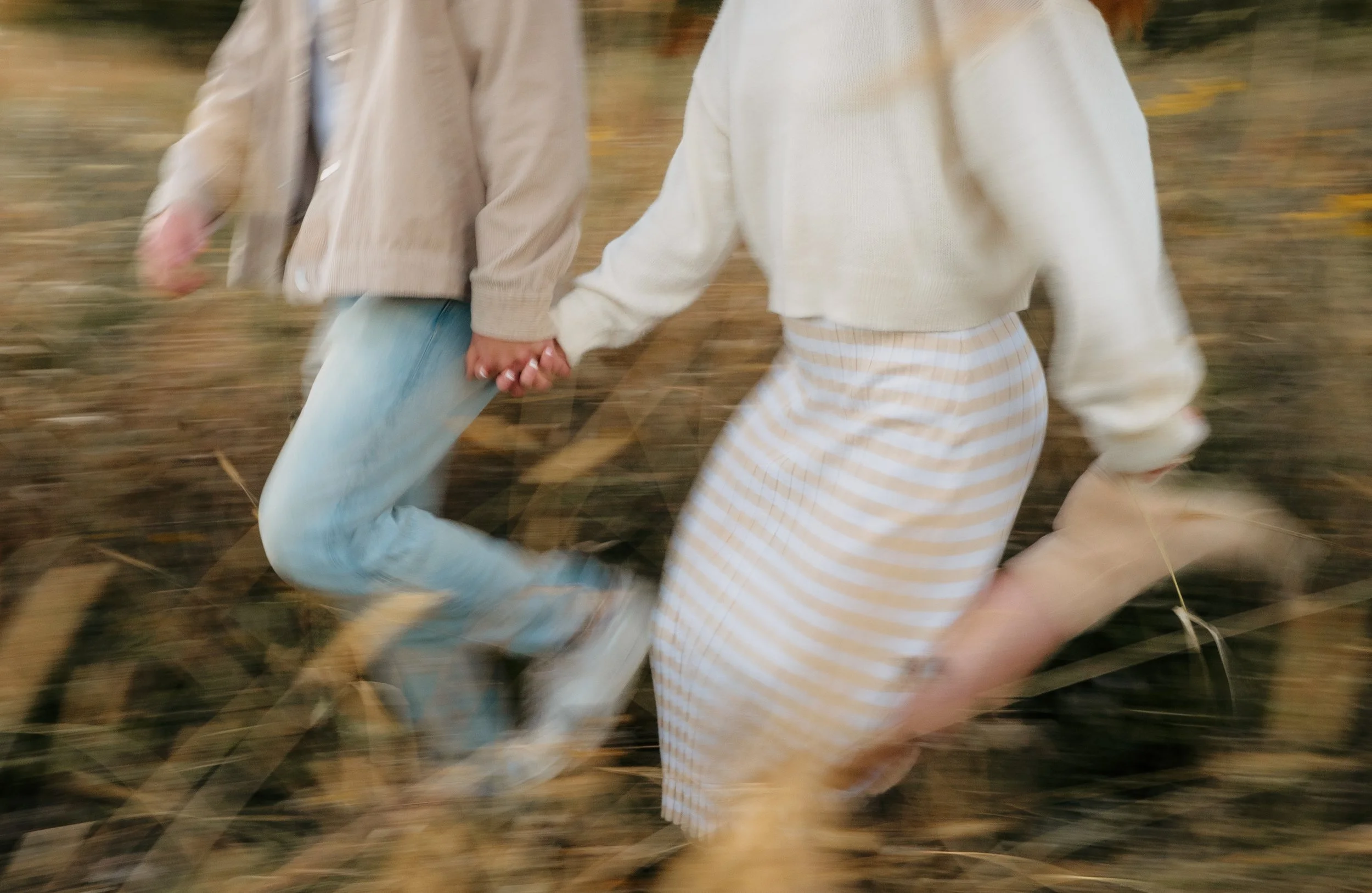 Two people holding hands while running through a field with blurred grass and leaves.