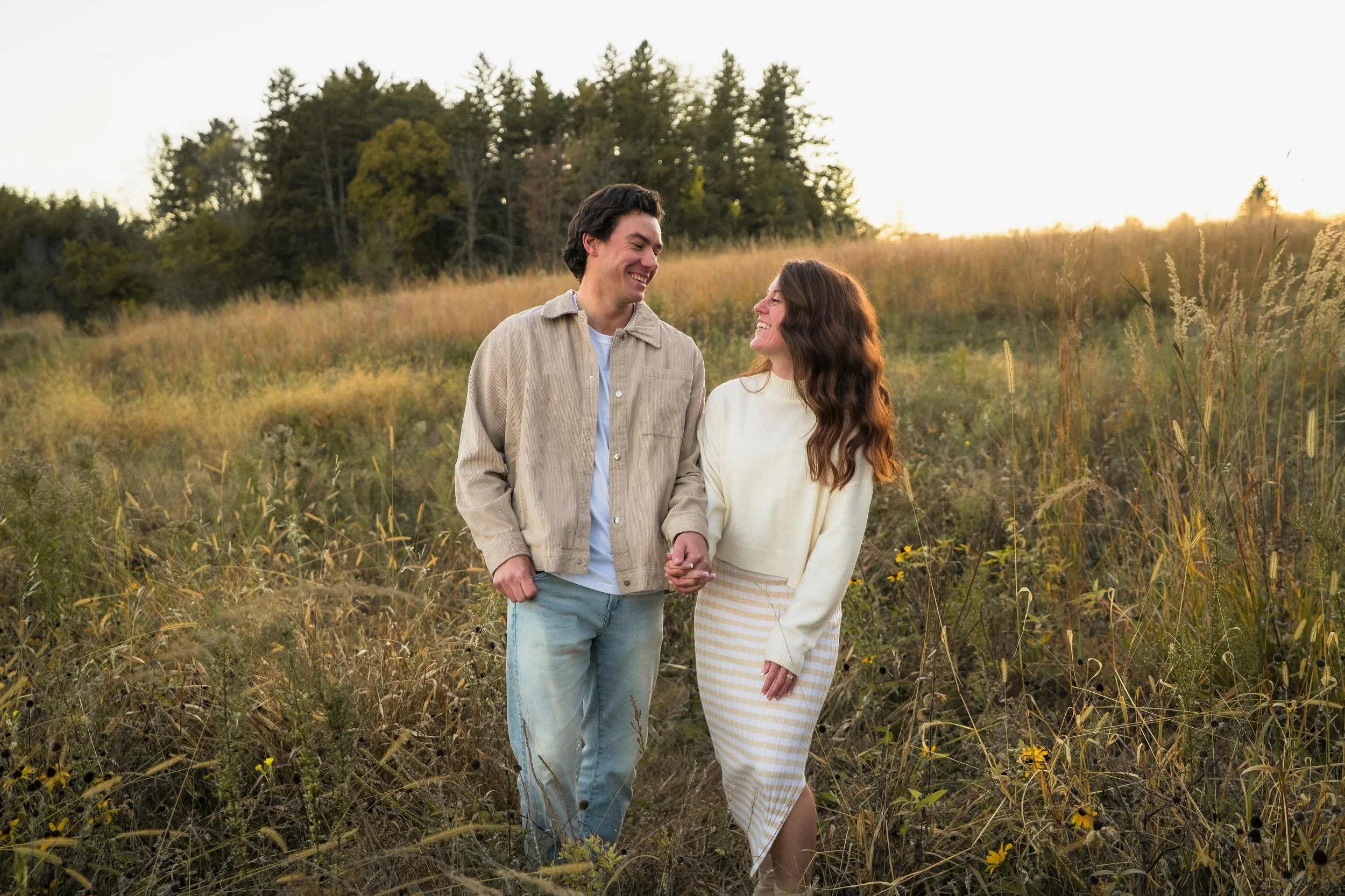 A young couple holding hands and smiling at each other while walking through a grassy field at sunset.