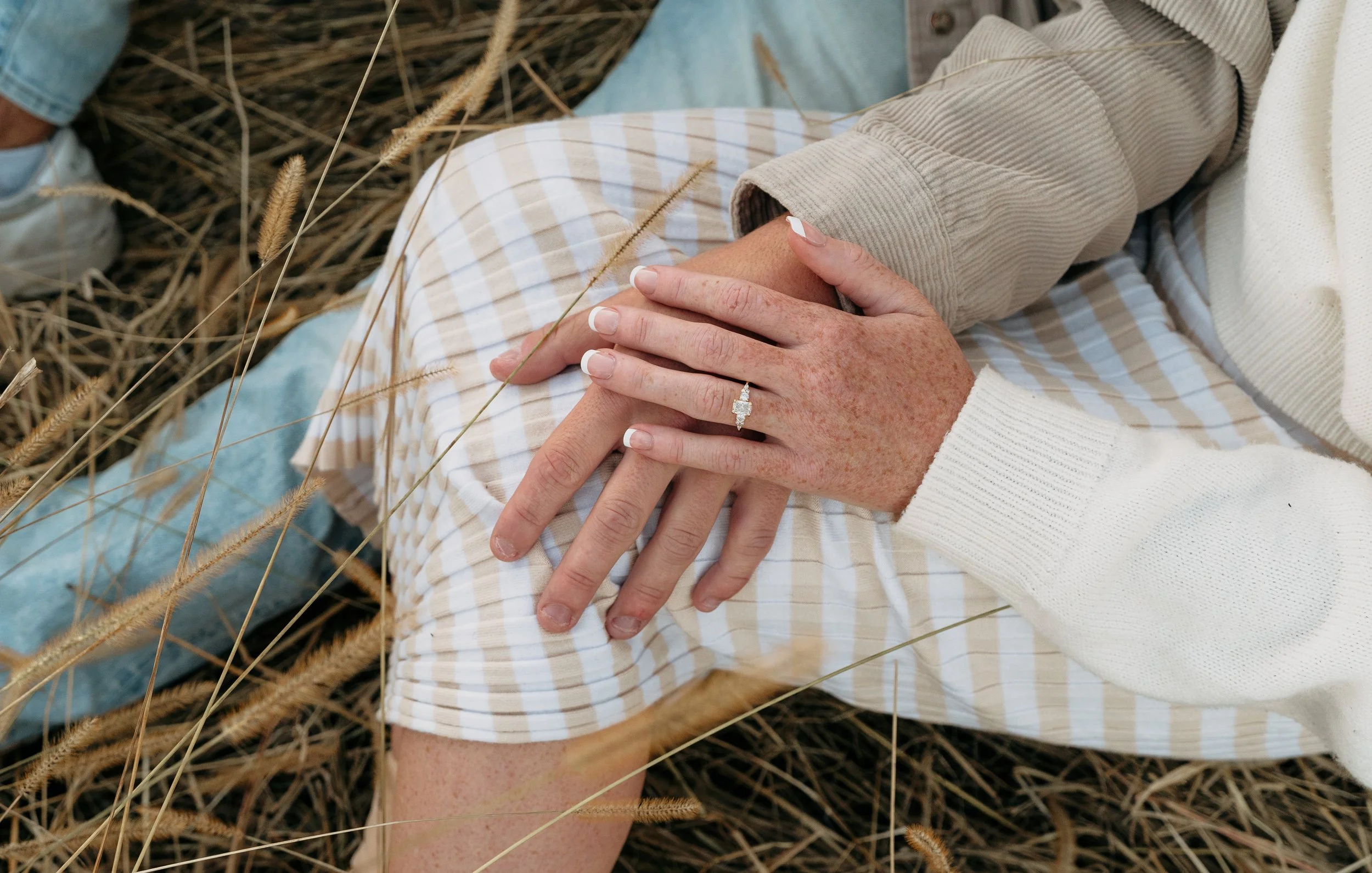 Close-up of a person's hands resting on their knee, showing a wedding ring, with grass and dried plants in the background. The person is wearing a striped skirt and a beige jacket.
