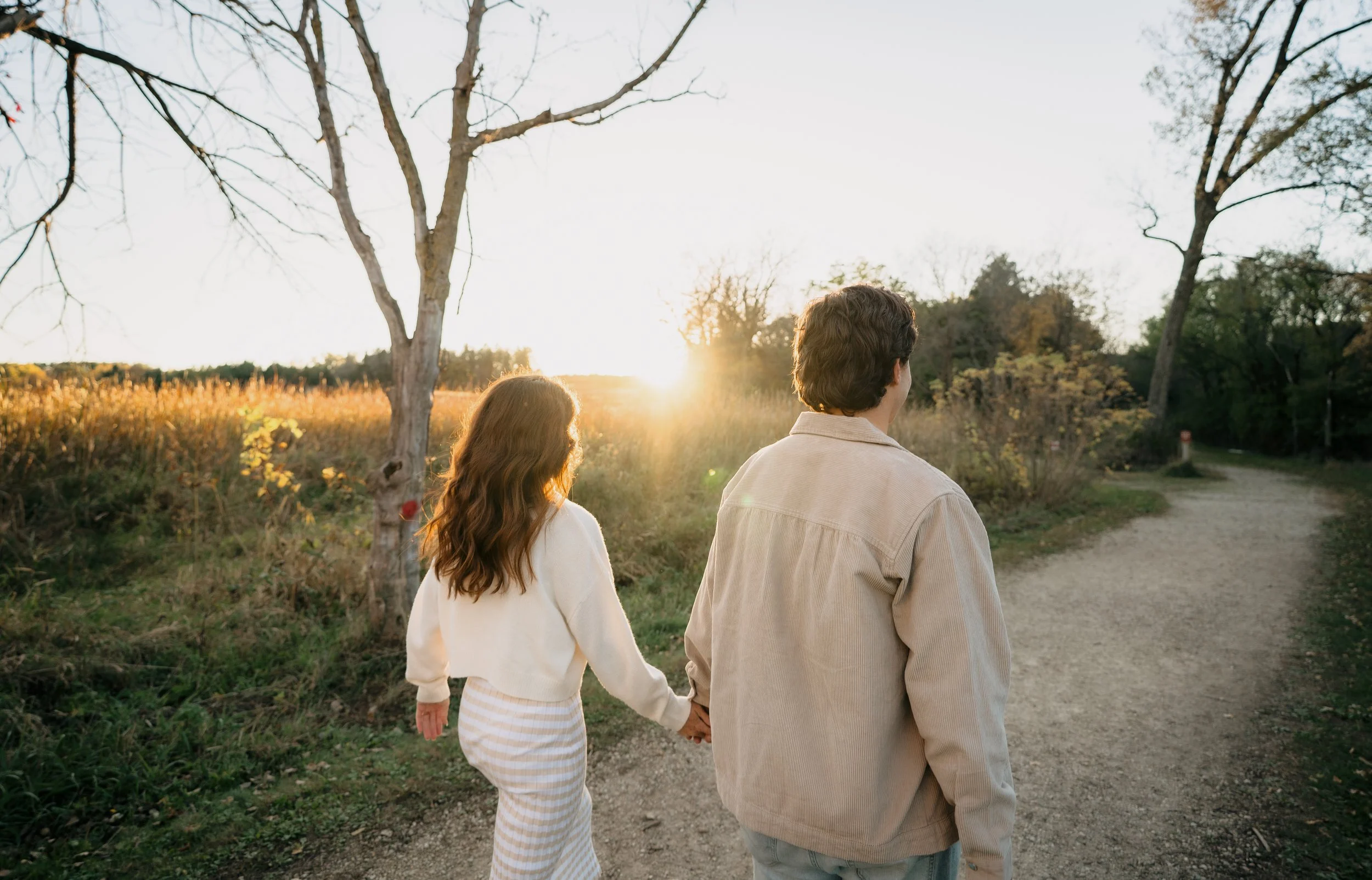 A man and woman walking hand in hand on a dirt path in a natural setting during sunset, surrounded by trees and grass.