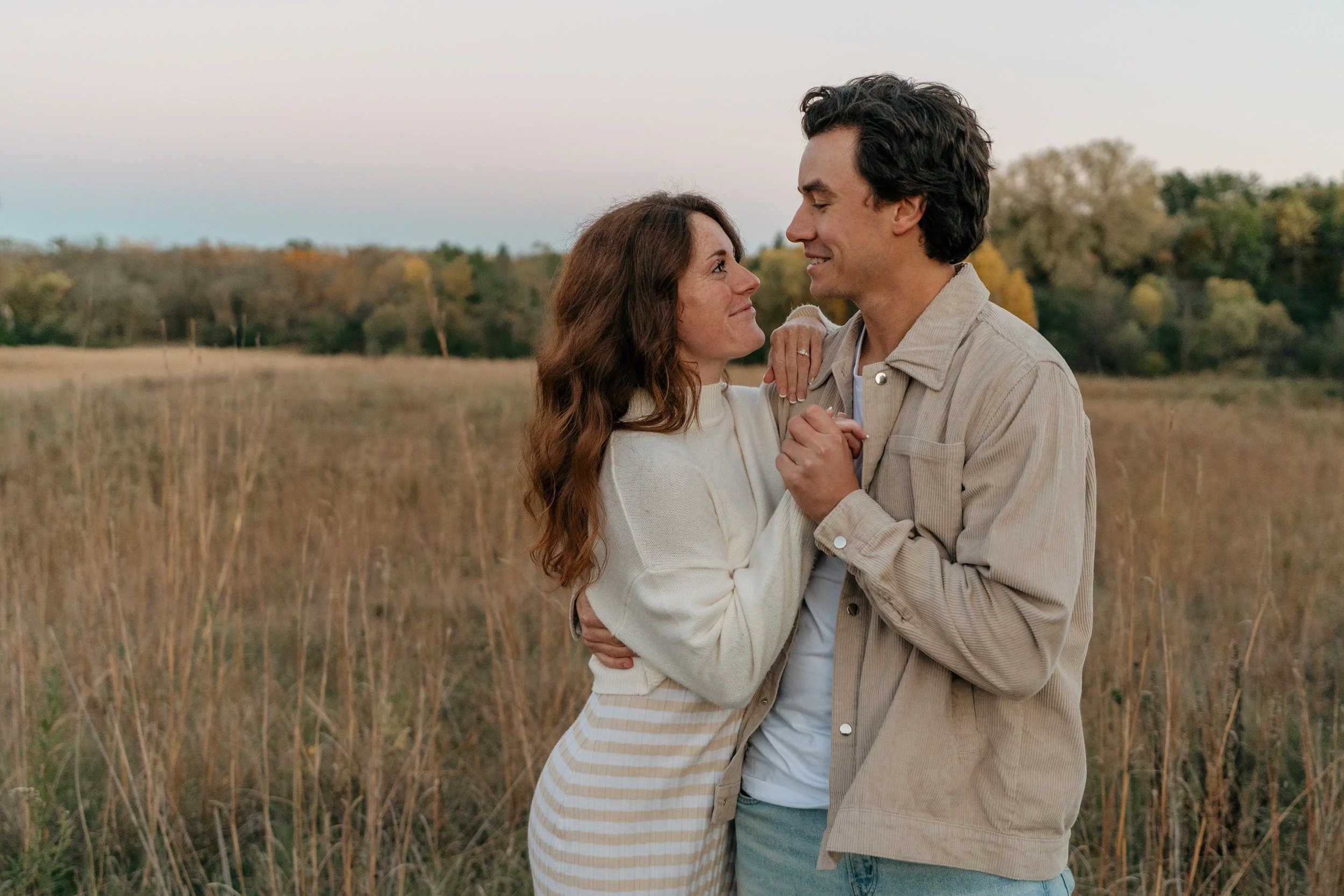 A couple standing in a field during sunset, looking lovingly at each other and holding hands.