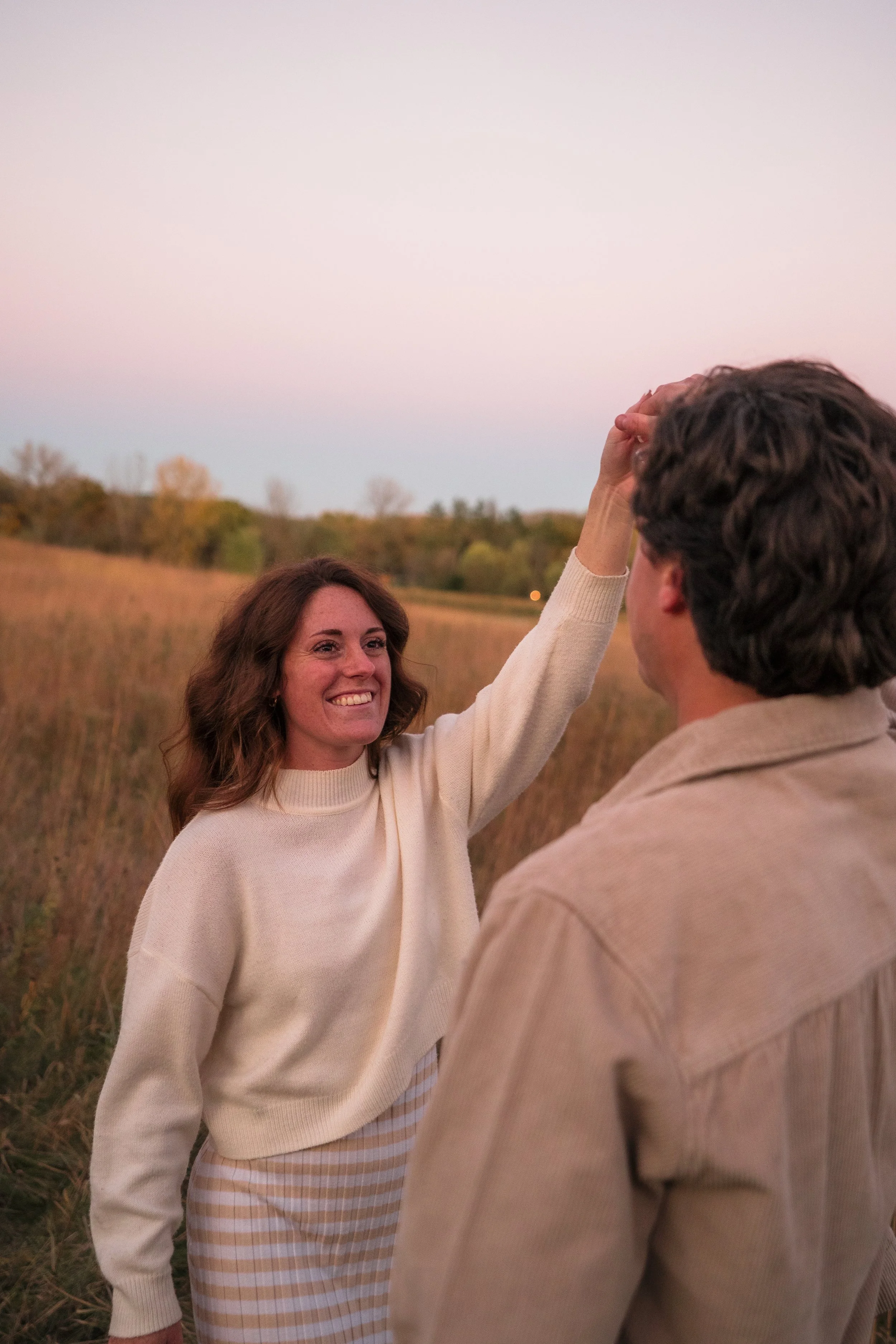 A woman and a man standing outdoors in a field during sunset. The woman is reaching up and touching the man's forehead while smiling. The woman has brown hair and is wearing a cream-colored sweater and a striped skirt. The man has curly hair and is w