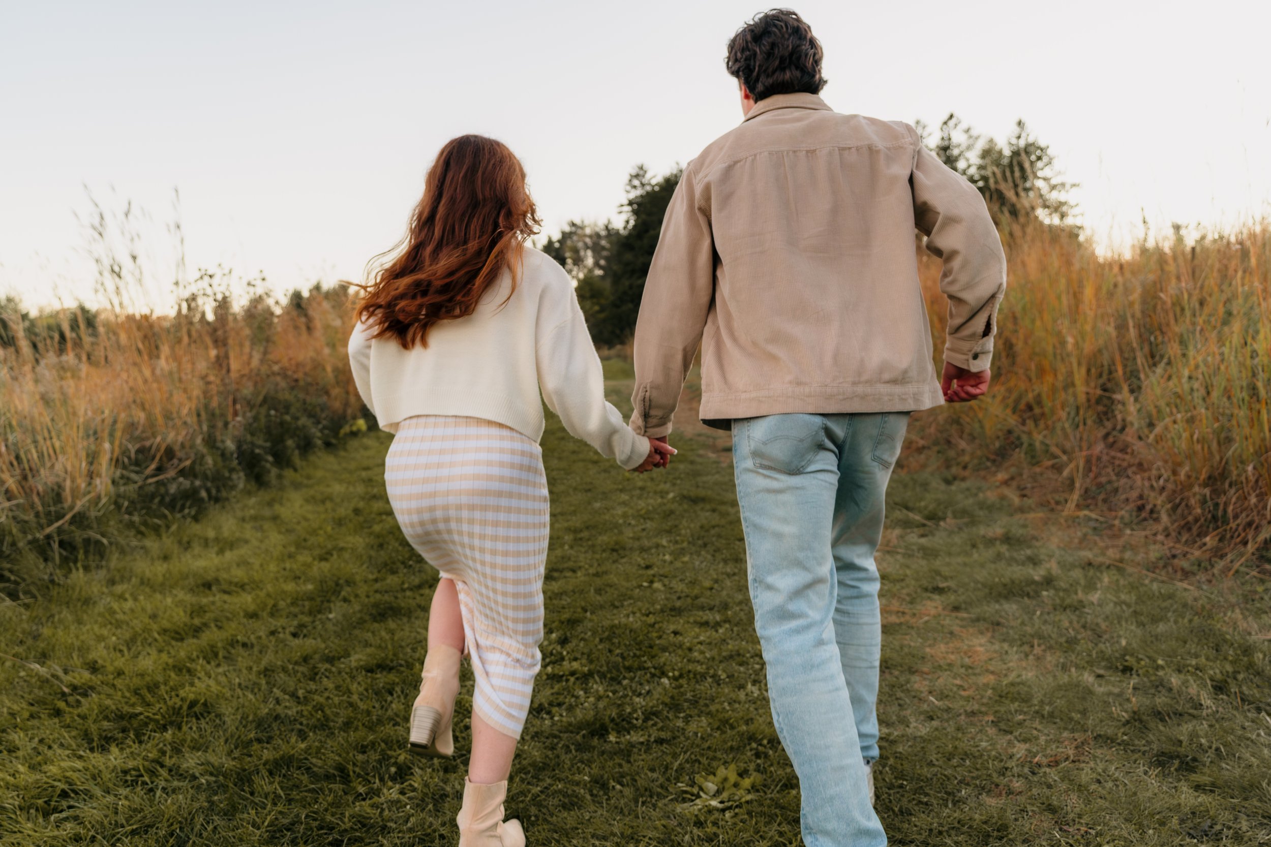 A man and woman holding hands walking on a grassy path in a field with tall grasses on either side during sunset.