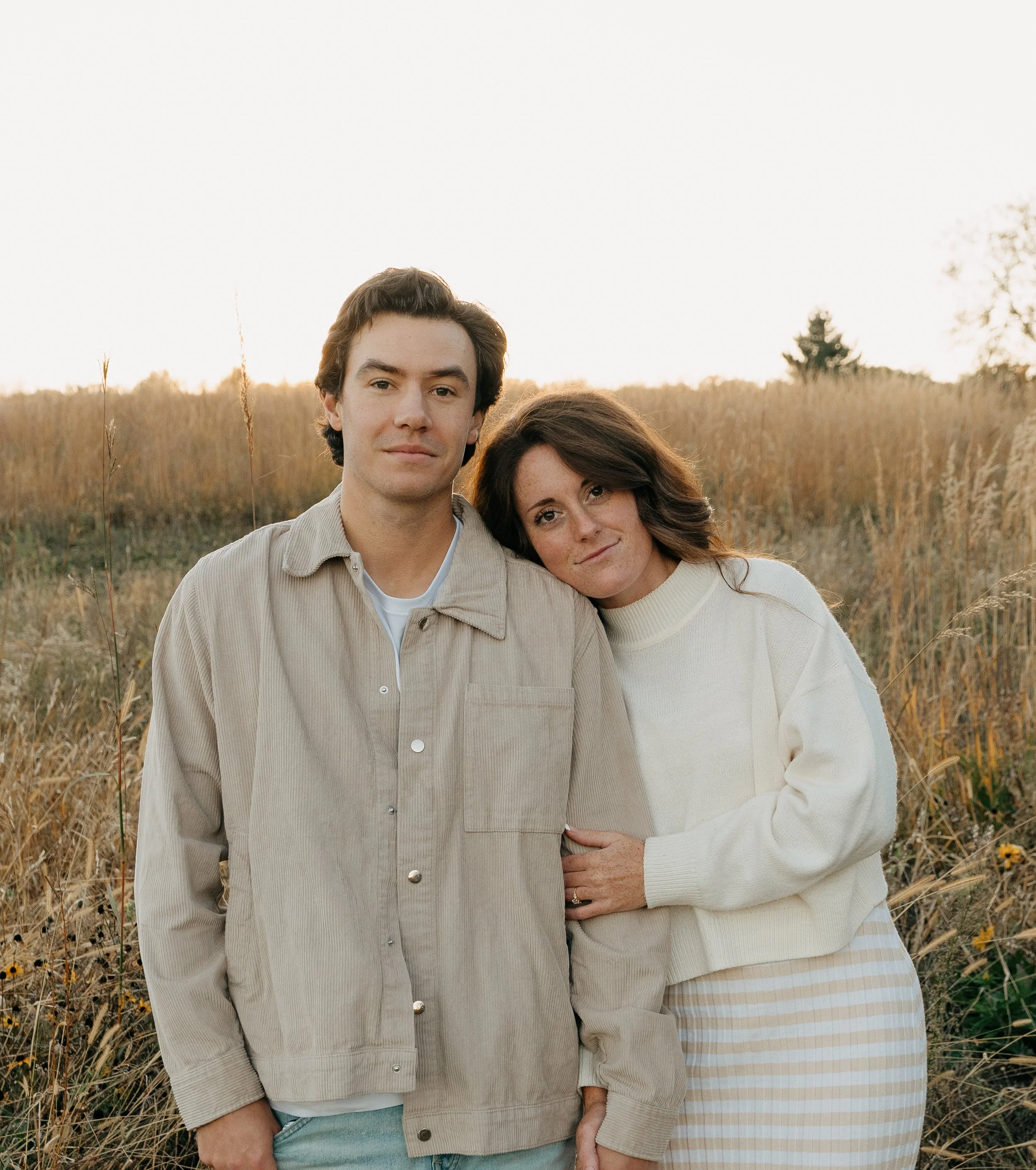 A young man and woman standing close together in a field of tall grass at sunset, with the woman leaning her head on the man's shoulder.