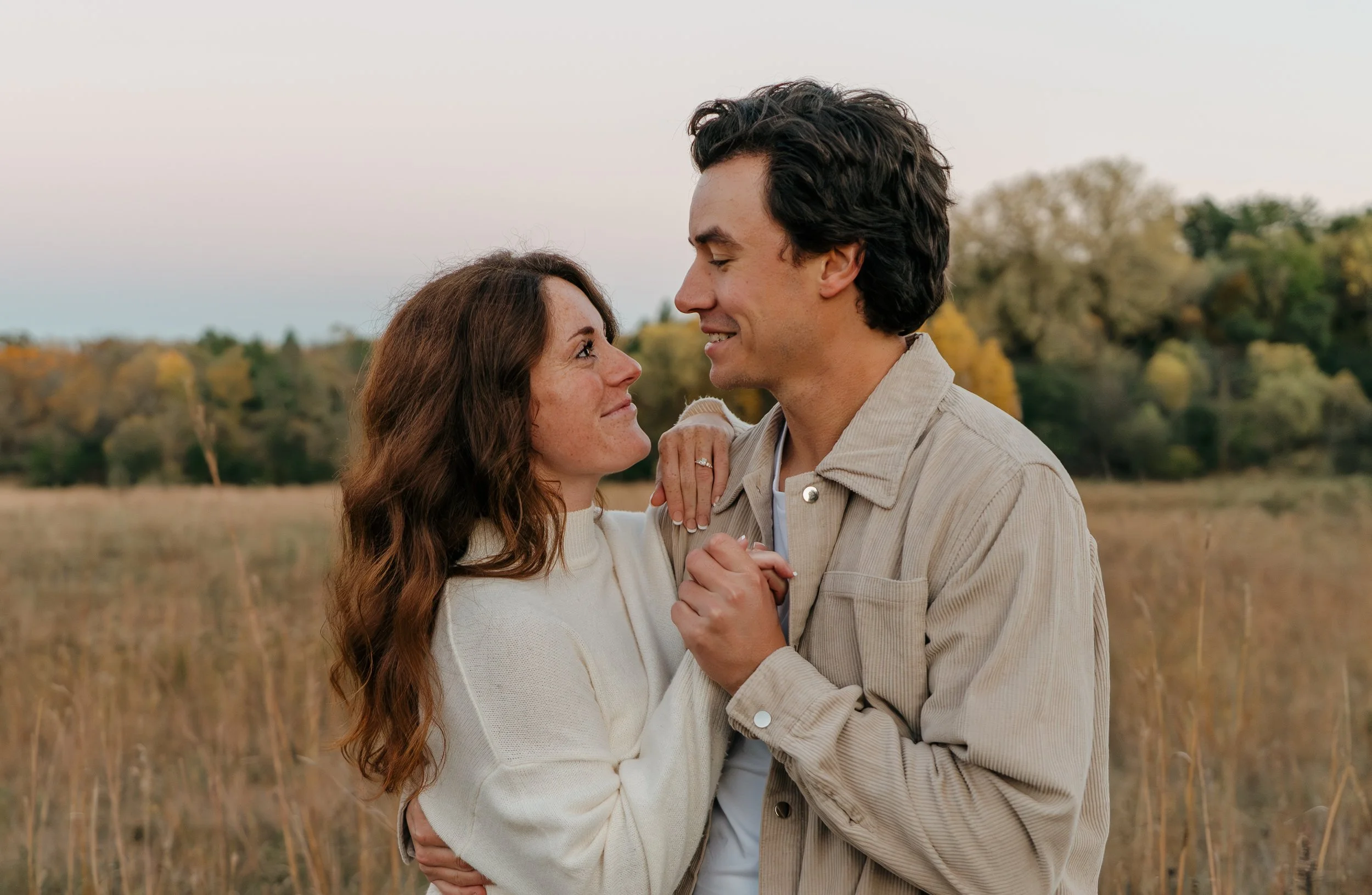 A couple standing close together outdoors in a field with trees in the background, smiling and looking into each other's eyes, with the woman's hand resting on the man's shoulder.