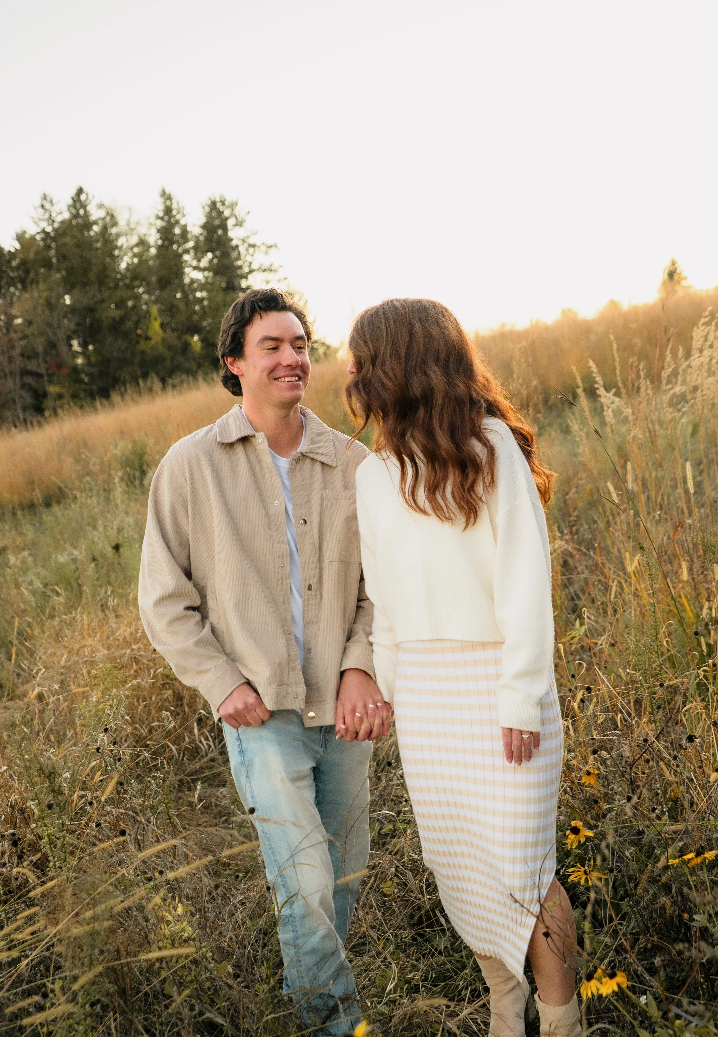 A young man and woman holding hands and walking through a field of tall grass and yellow flowers during sunset, smiling at each other.