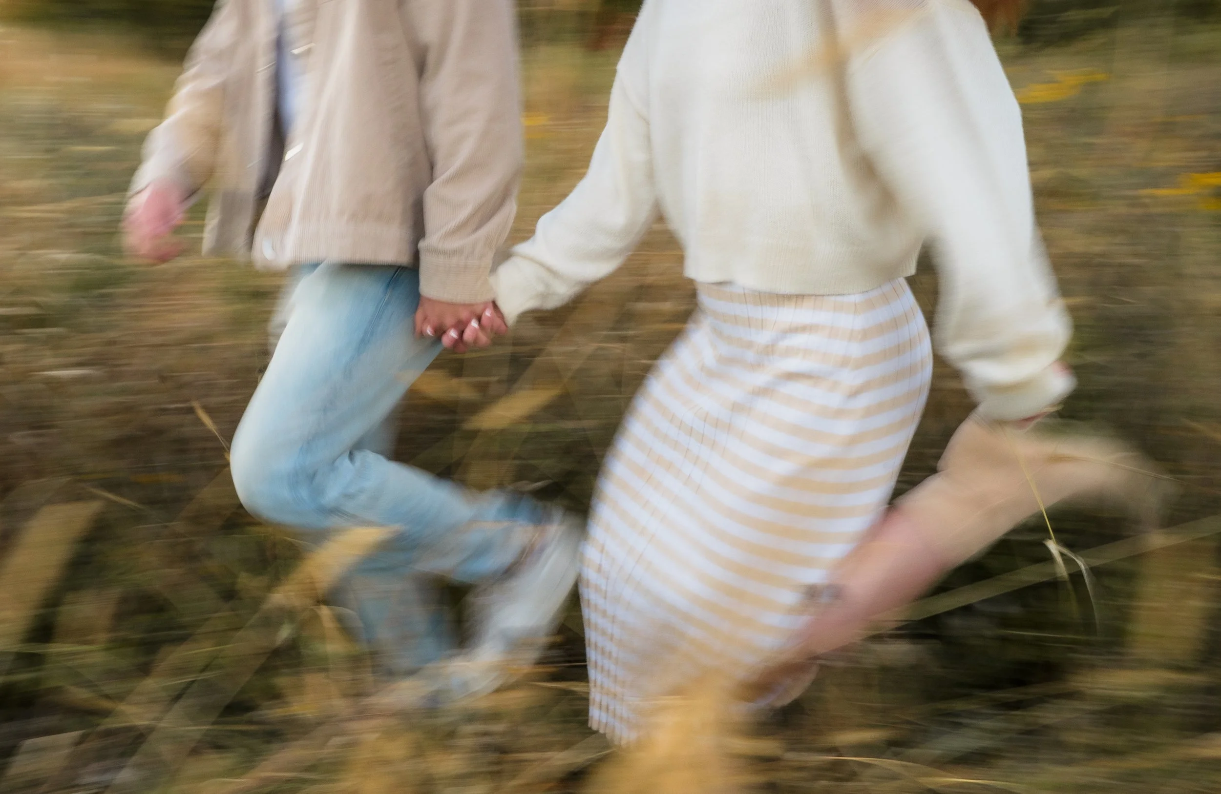 Two people holding hands while running through an outdoor field with tall grass and yellow flowers in the background.