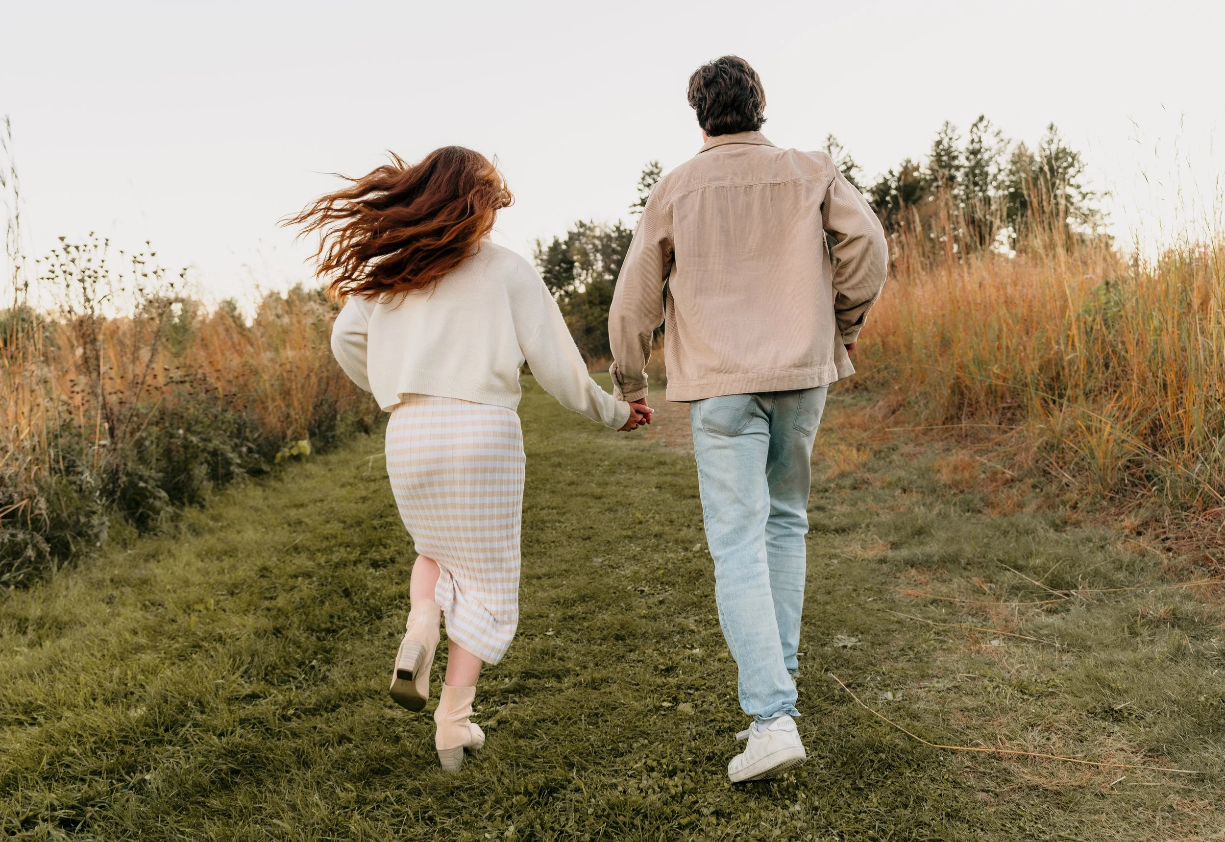 A couple holding hands walking on a grassy path in a field during autumn, with tall dry grass and trees in the background.