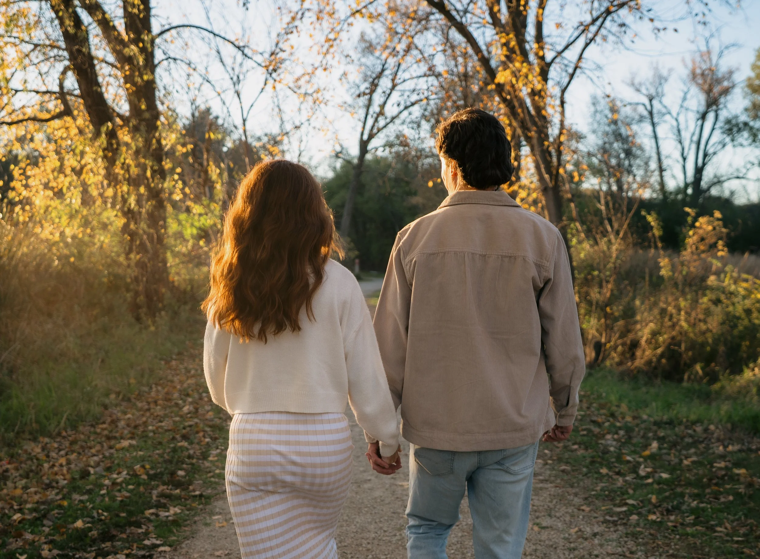 A man and woman walking hand in hand on a dirt path in a park during autumn, with trees and fallen leaves around them, illuminated by warm sunlight.