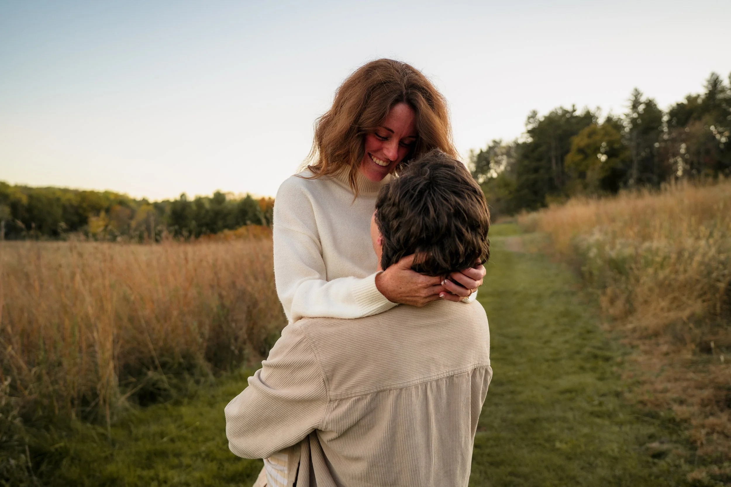 A woman lifts a man in a field, smiling at each other, with trees in the background at sunset.