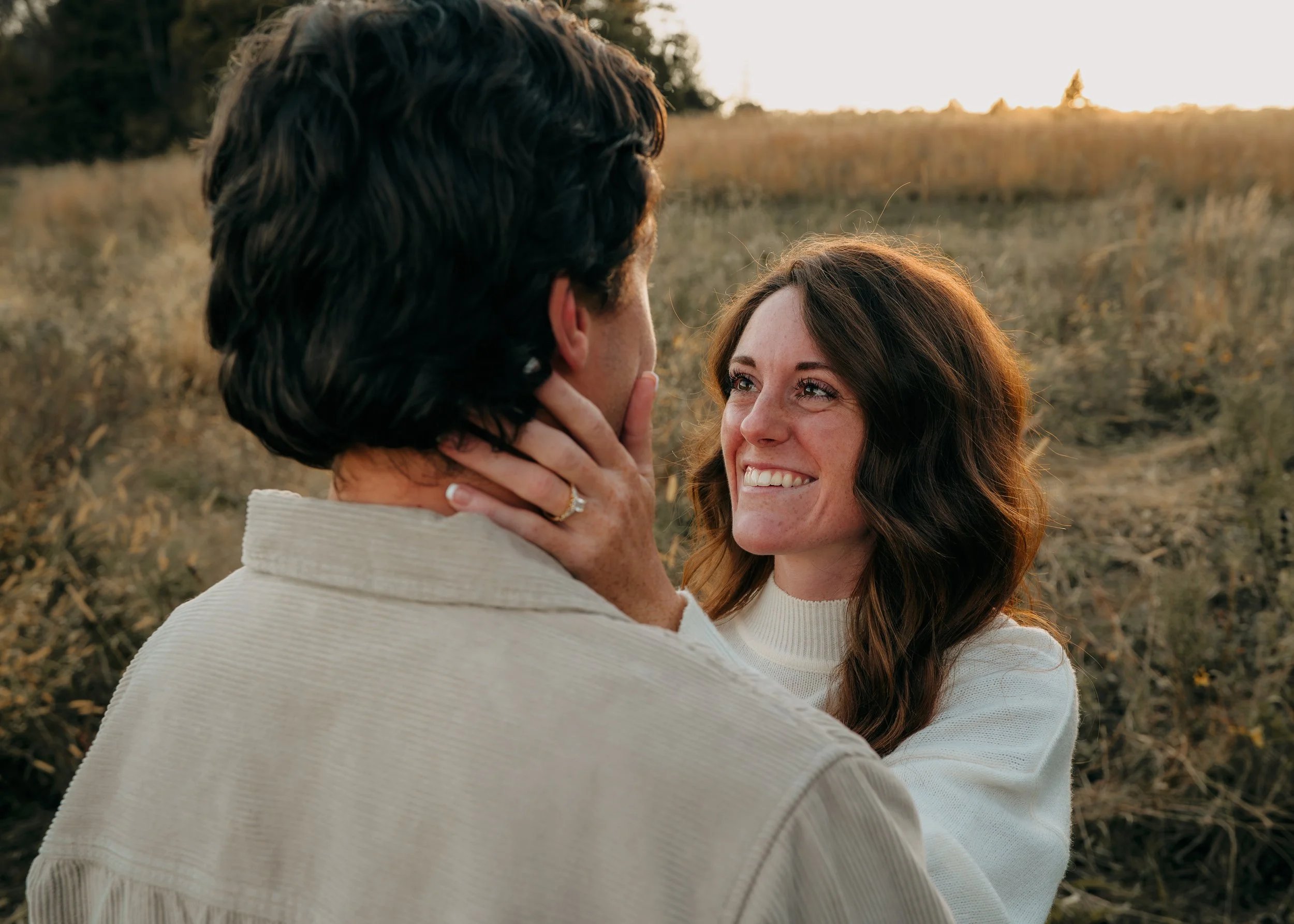 A woman with long brown hair smiling and touching a man's face outdoors at sunset, standing in a field of tall grass.