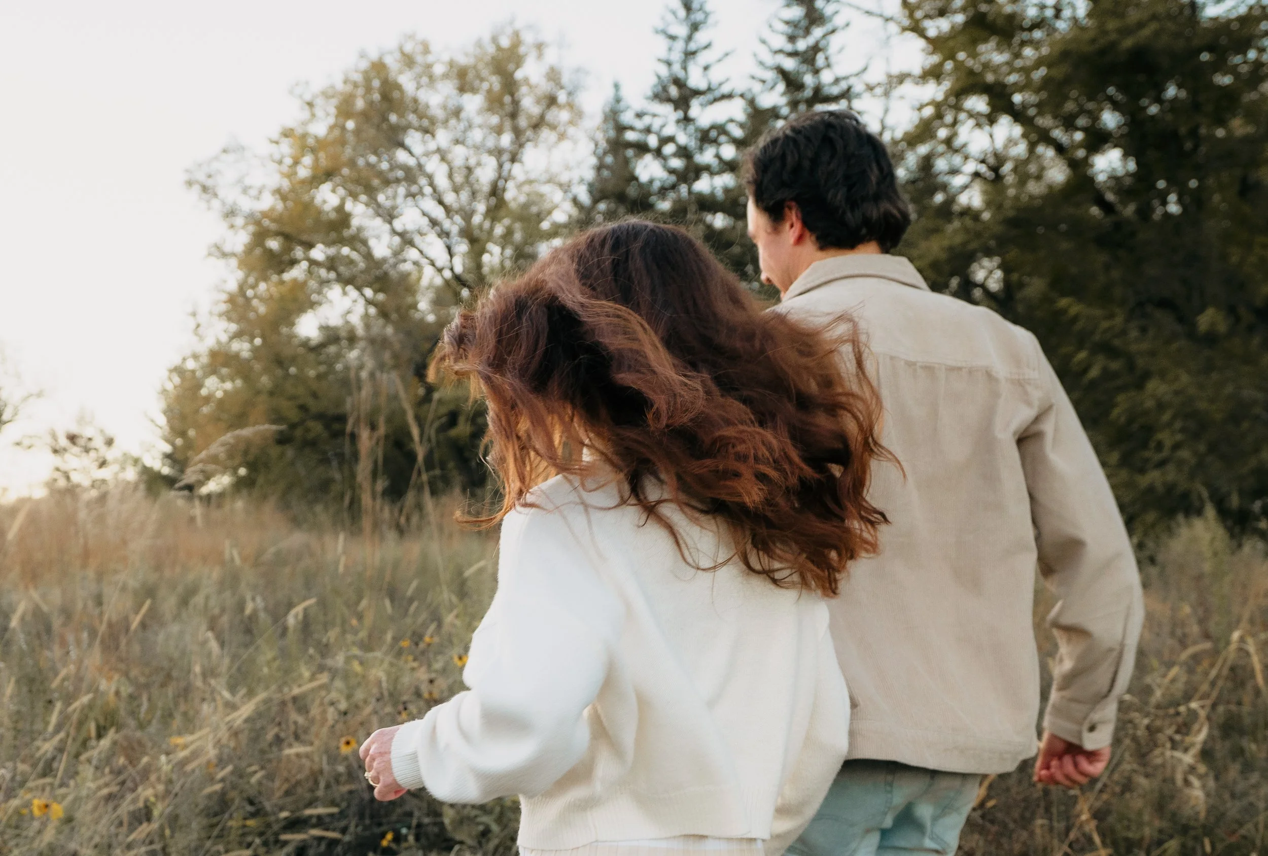A couple walking outdoors through a field of tall grass and wildflowers, with trees and a pale sky in the background.