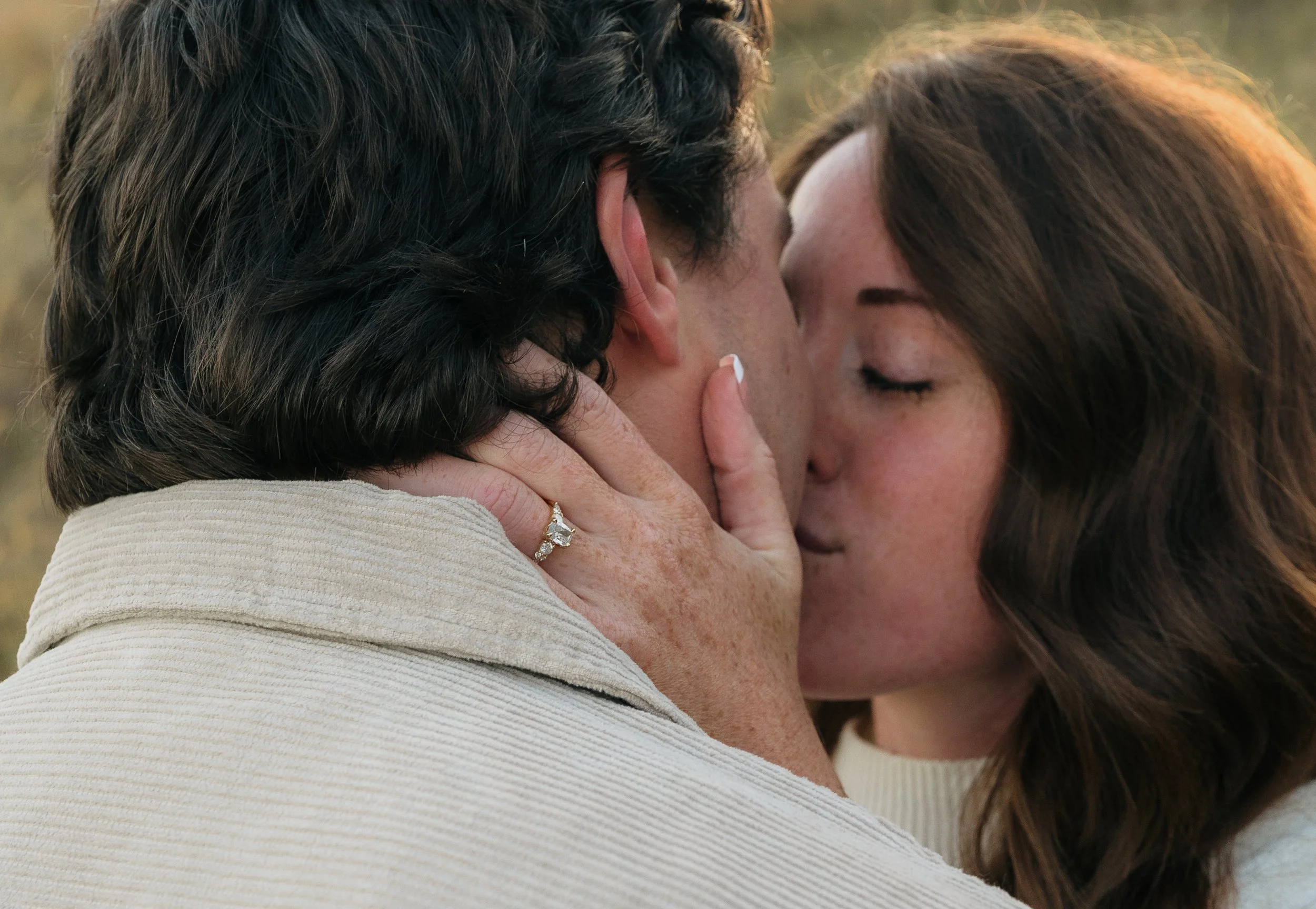 A couple sharing a kiss outdoors, the woman has dark brown hair and is wearing an engagement ring. The man has dark hair and is wearing a light-colored shirt.