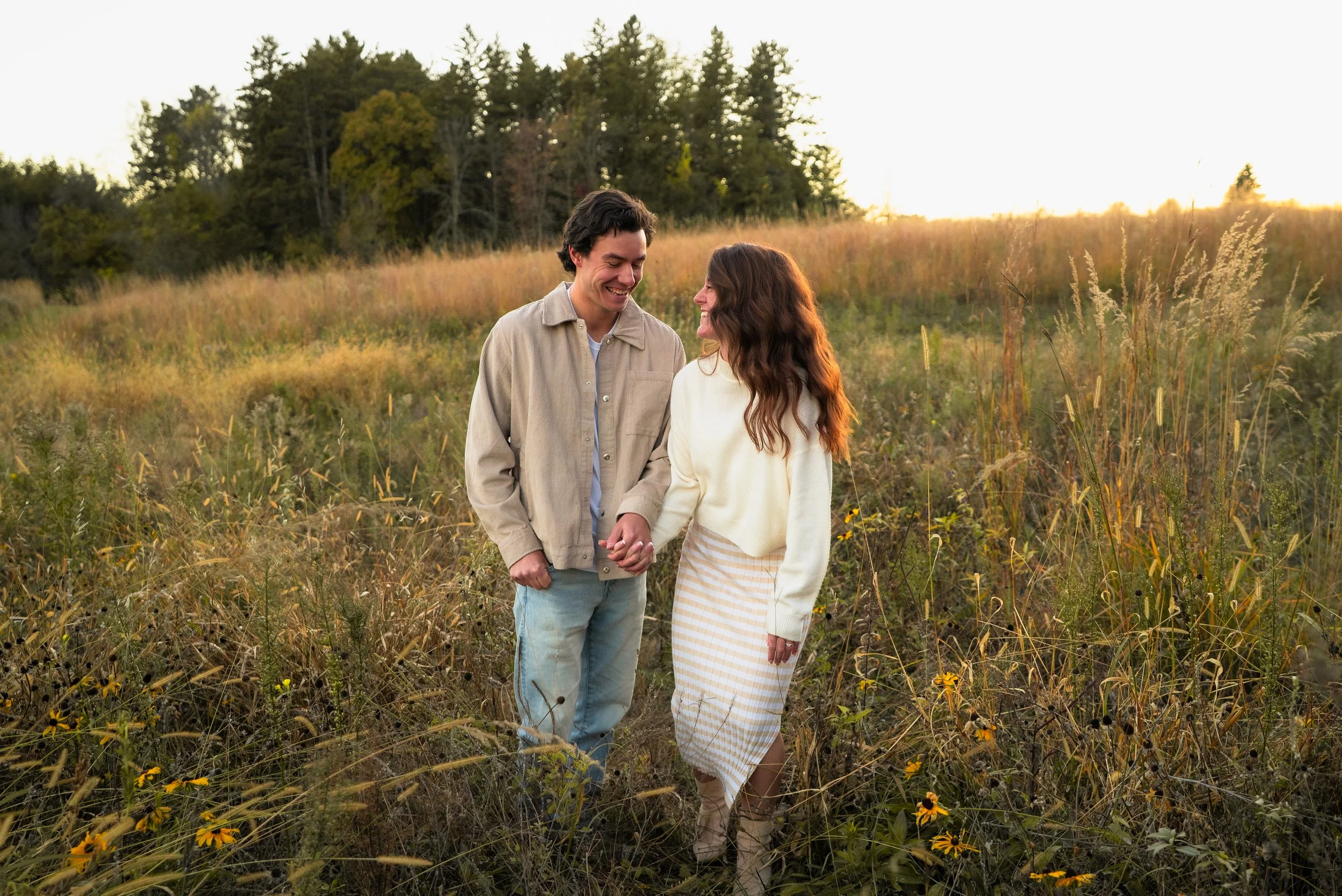 A young couple holding hands and smiling in a grassy field during sunset with trees in the background.