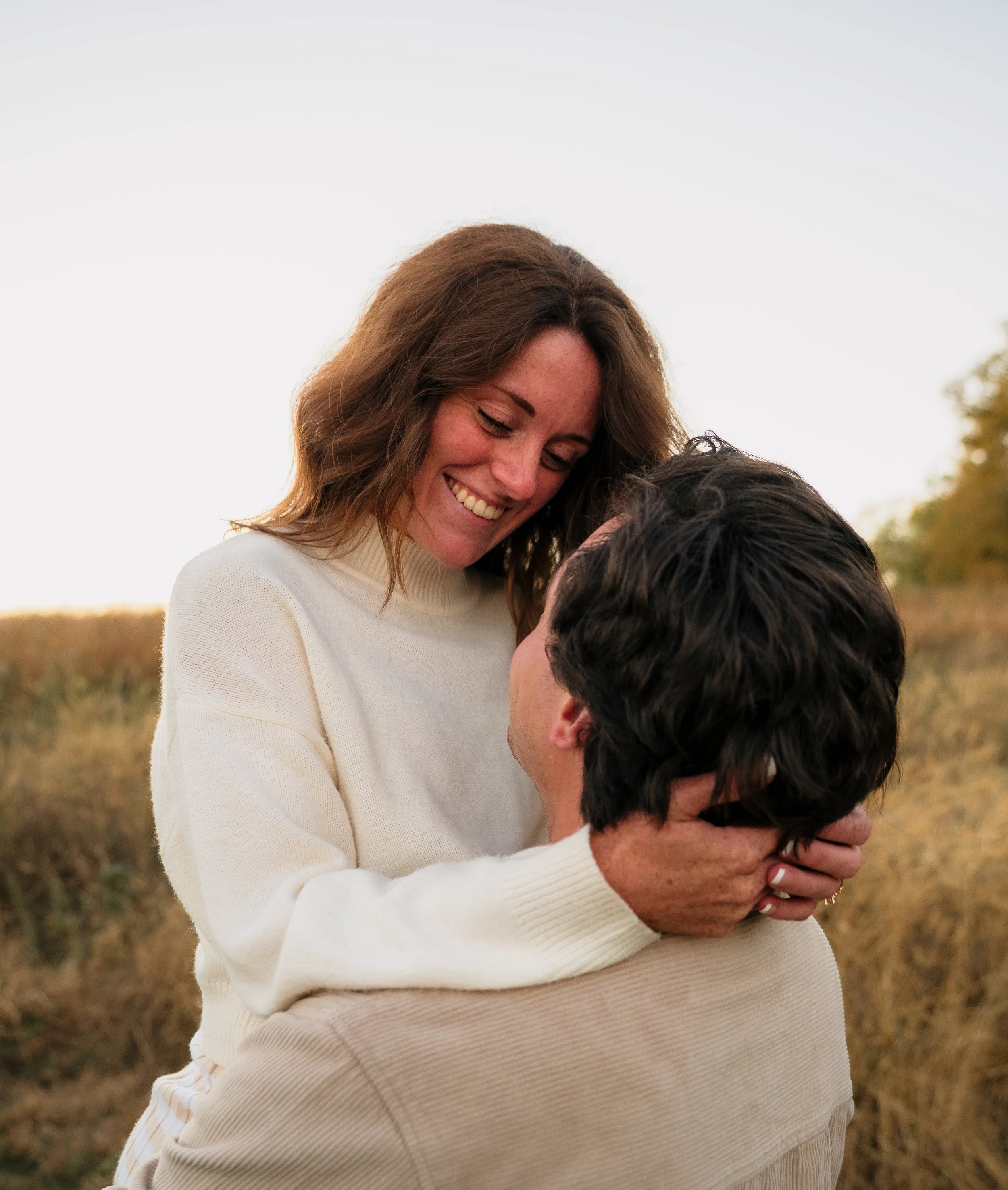 A smiling woman with brown hair holding a man with dark hair in an outdoor setting with a field and trees in the background.