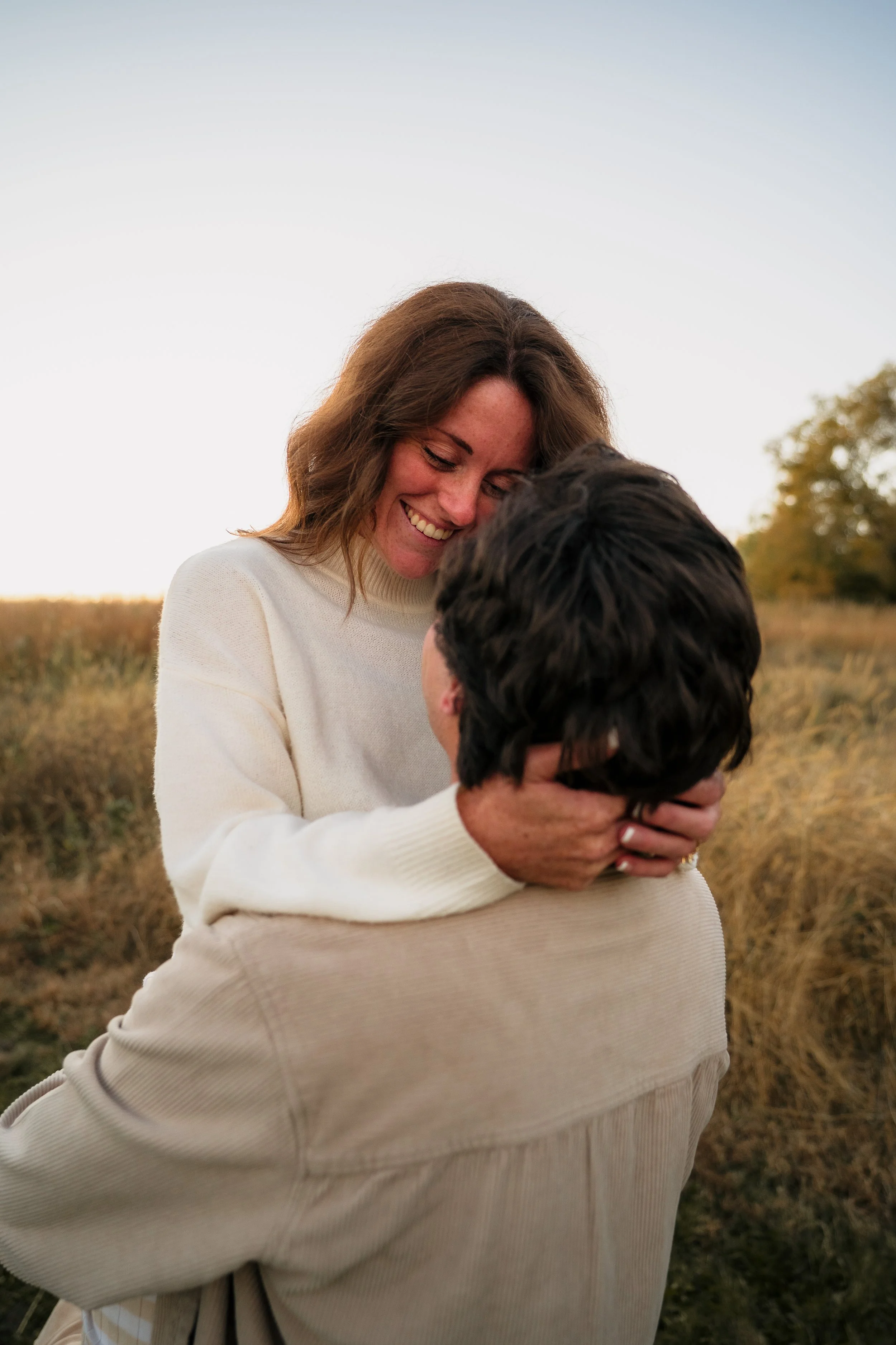 A woman with wavy brown hair and a woman with short black hair sharing a hug in a field at sunset.