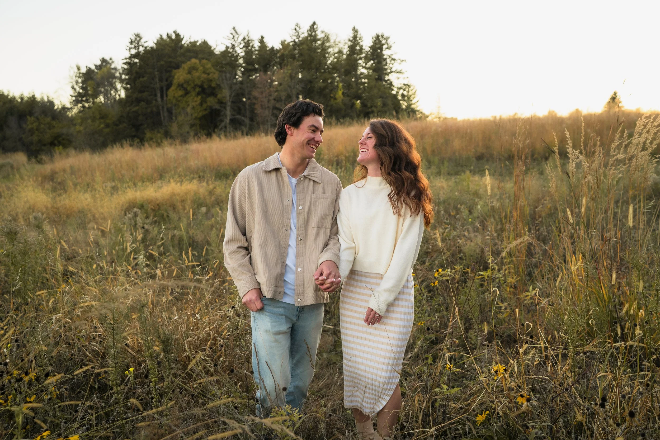 A smiling couple holding hands and walking through a grassy field during sunset.