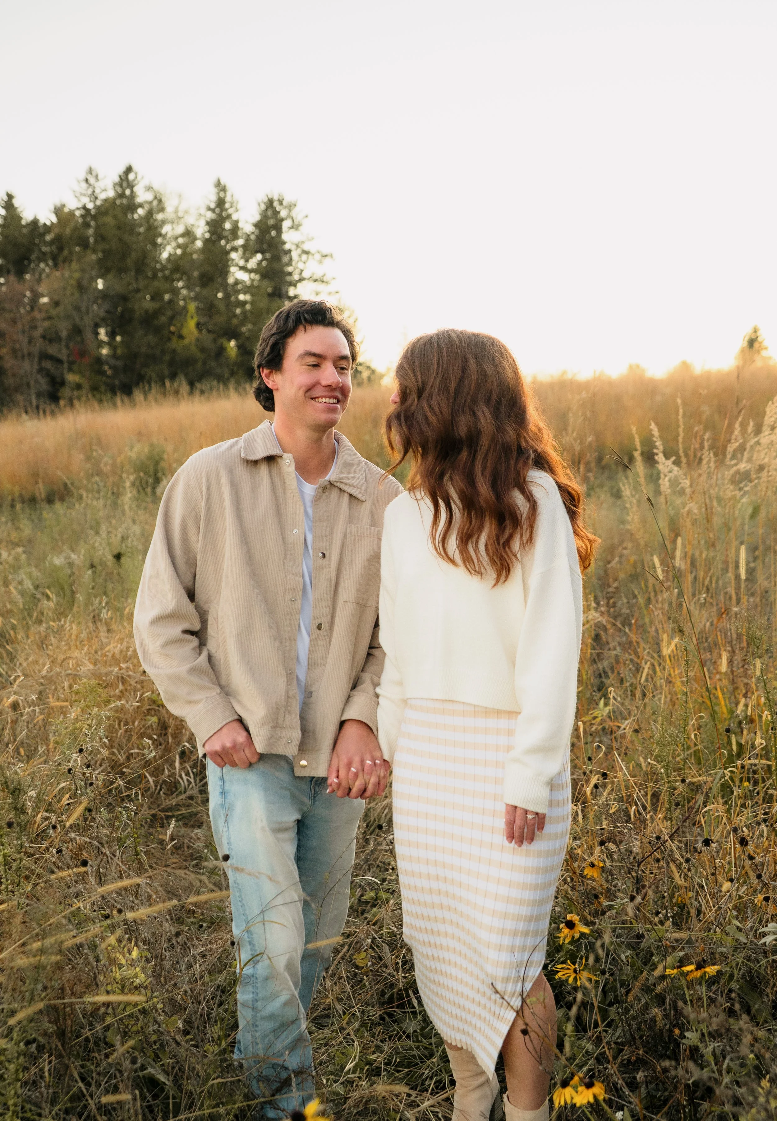 A couple holding hands and smiling at each other in a field during sunset.